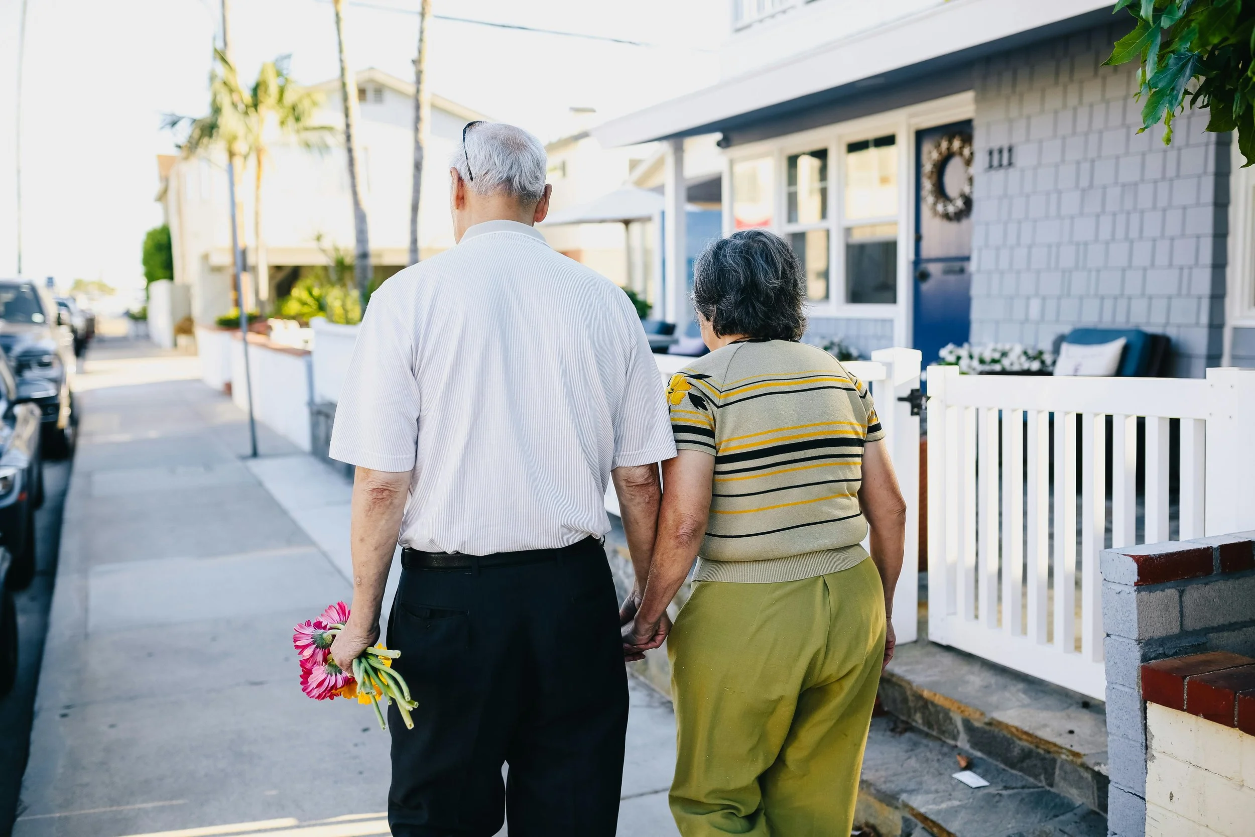 An elderly couple walking down a sidewalk, holding hands, with the man holding a small bouquet of pink flowers in his left hand.