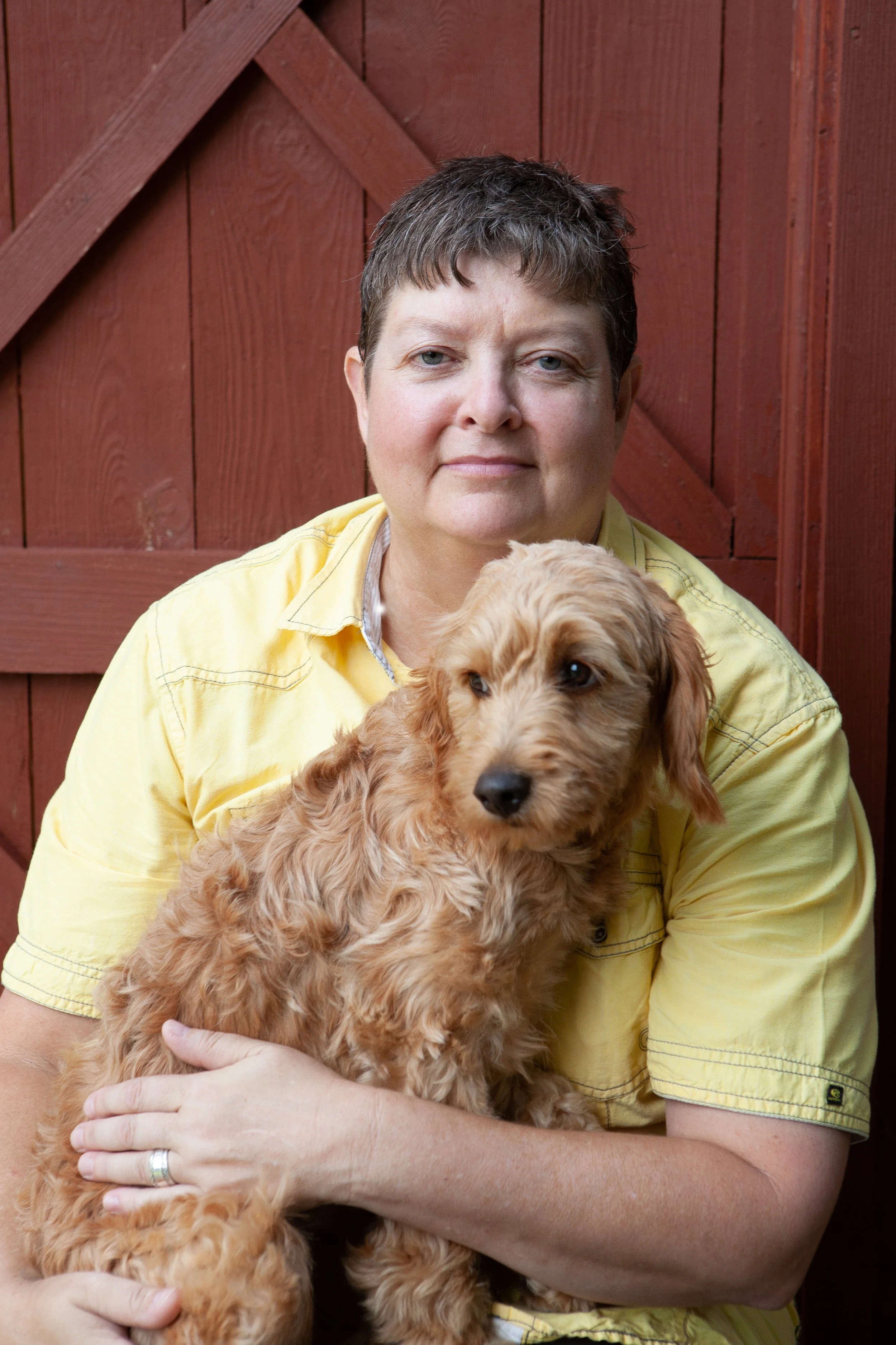 A woman with short hair wearing a yellow shirt holding a tan, curly-haired dog in front of a red wooden fence.