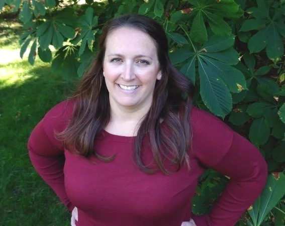 A woman with long brown hair smiling in front of large green leaves, wearing a maroon long-sleeve shirt.