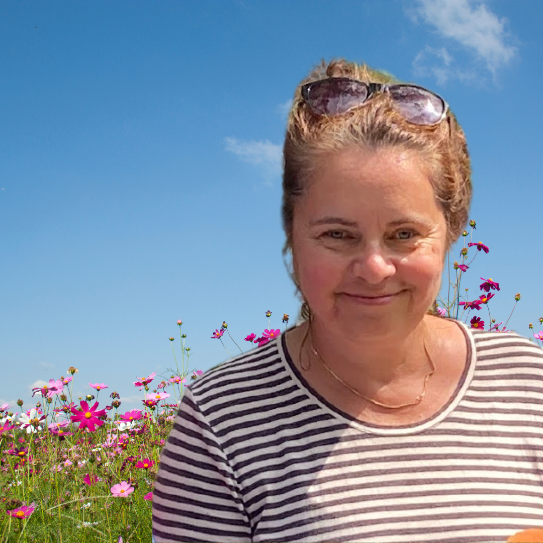 A woman with blonde hair, wearing sunglasses on her head and a striped shirt, smiling in front of a field of pink and purple flowers under a clear blue sky.