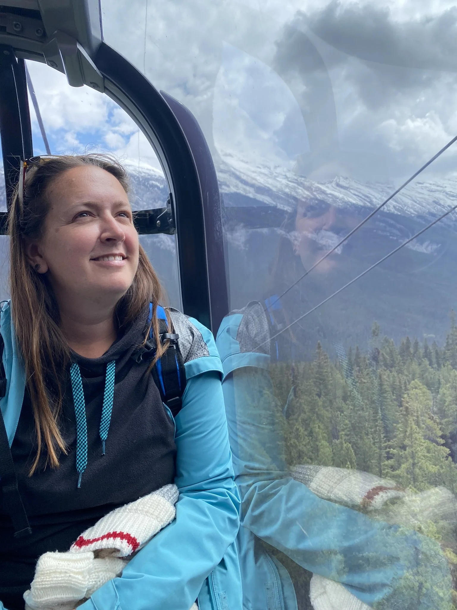 Woman sitting inside a cable car with a mountain and forest view reflected on the glass behind her.