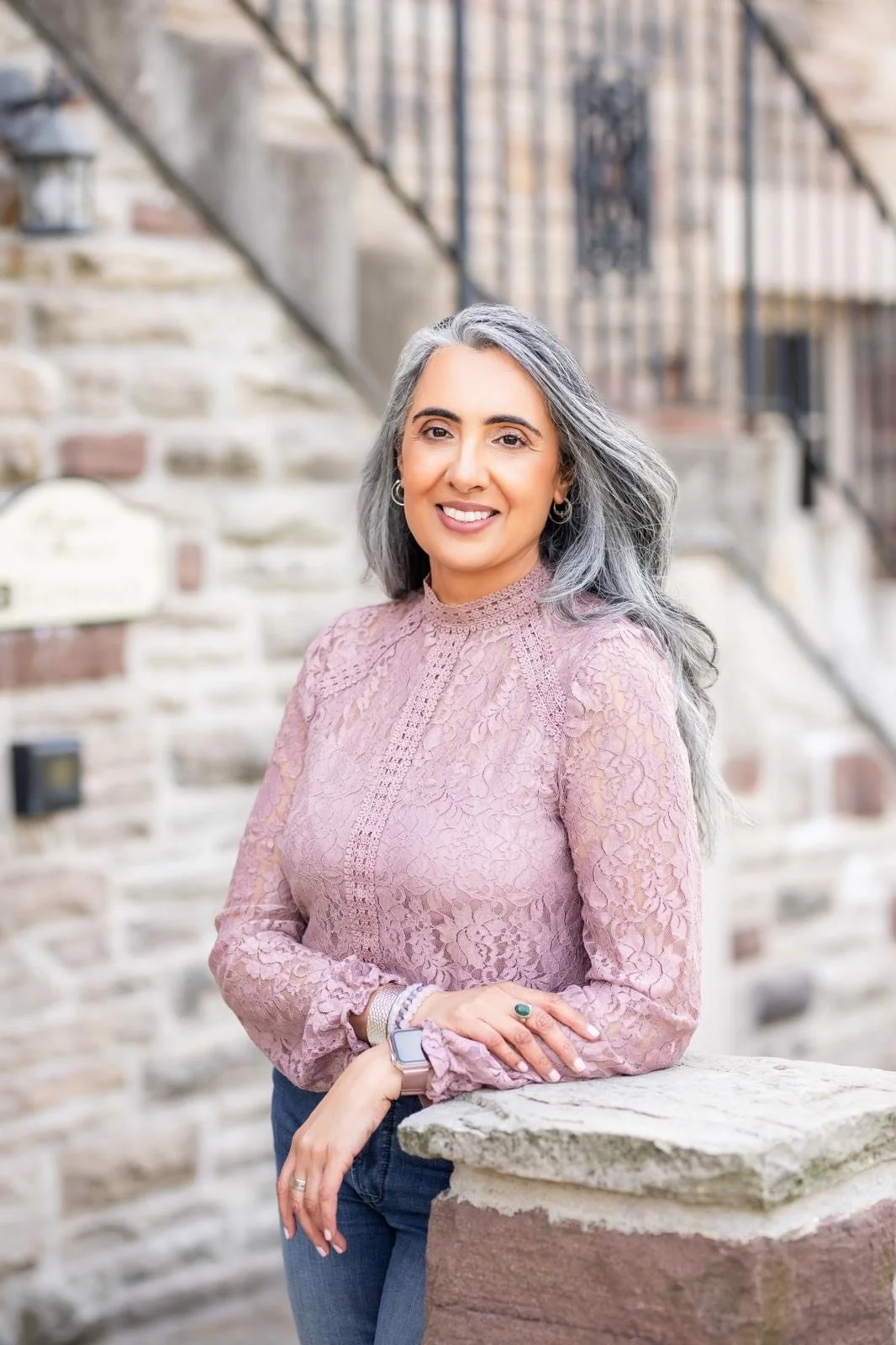 A woman with gray hair, wearing a pink lace top and blue jeans, smiling and standing outdoors with a stone wall and stairs in the background.