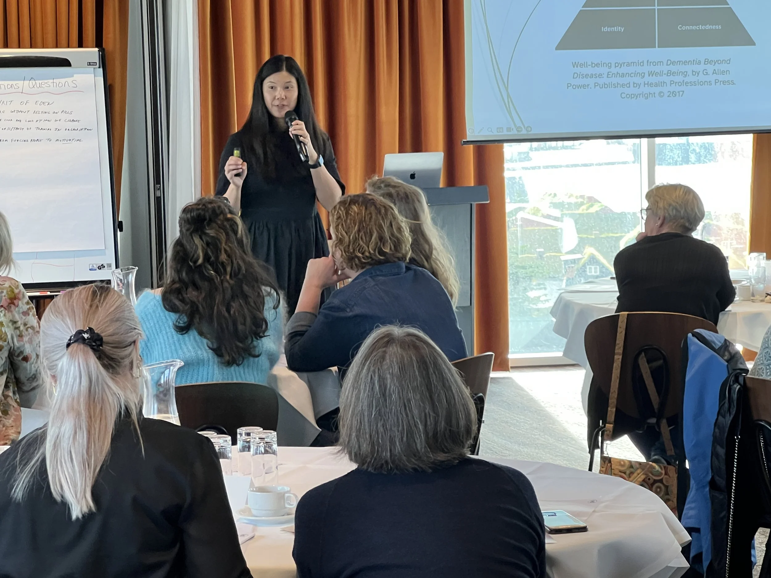 A woman in a black dress giving a presentation to a group of adults in a conference room. She holds a microphone and a remote, standing in front of a large screen displaying a PowerPoint slide, with some attendees taking notes or listening attentively.