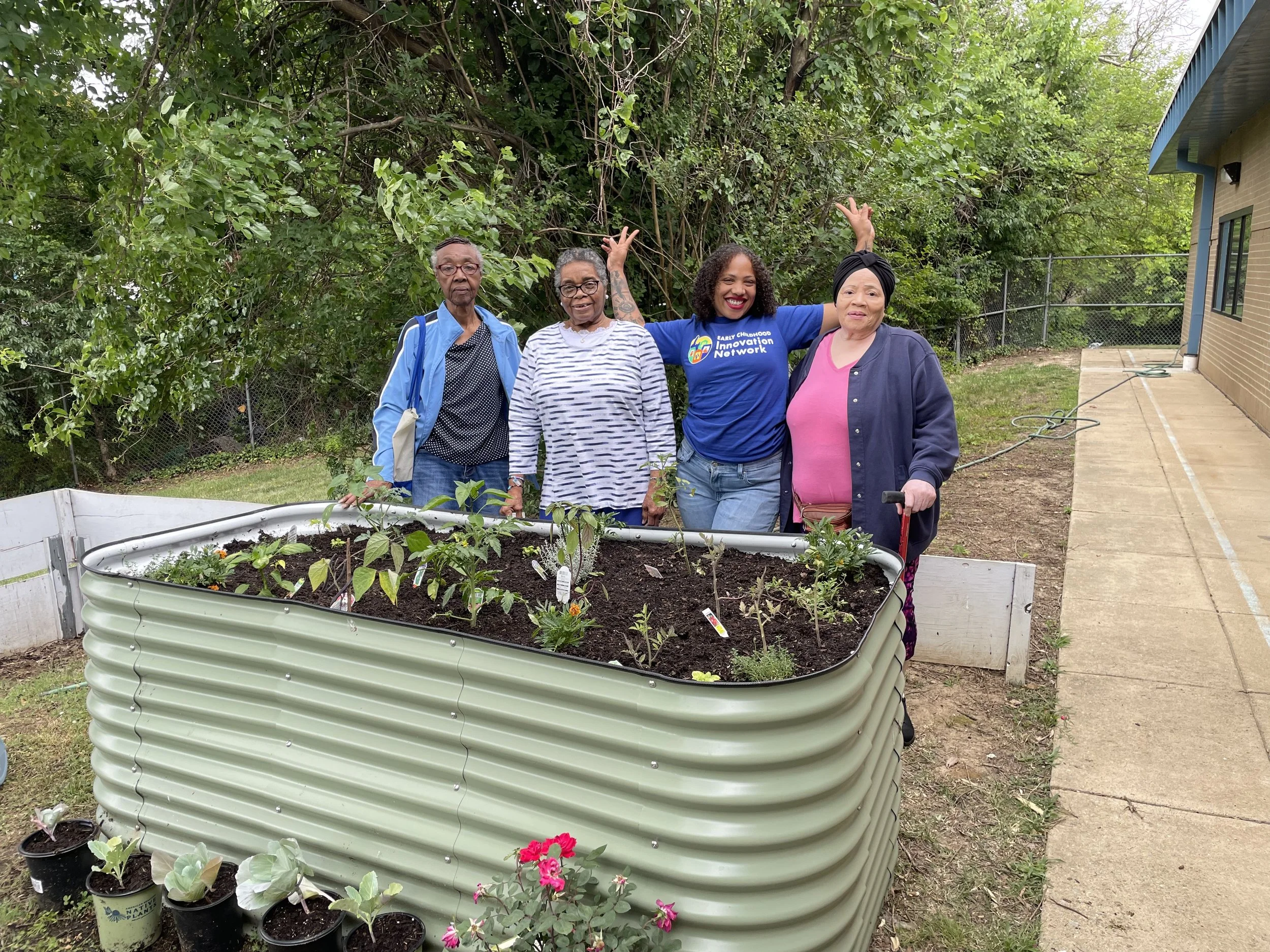 Four women stand in a garden near a raised garden bed filled with young plants. They are smiling and posing together, with a large tree and a house with a blue roof in the background.
