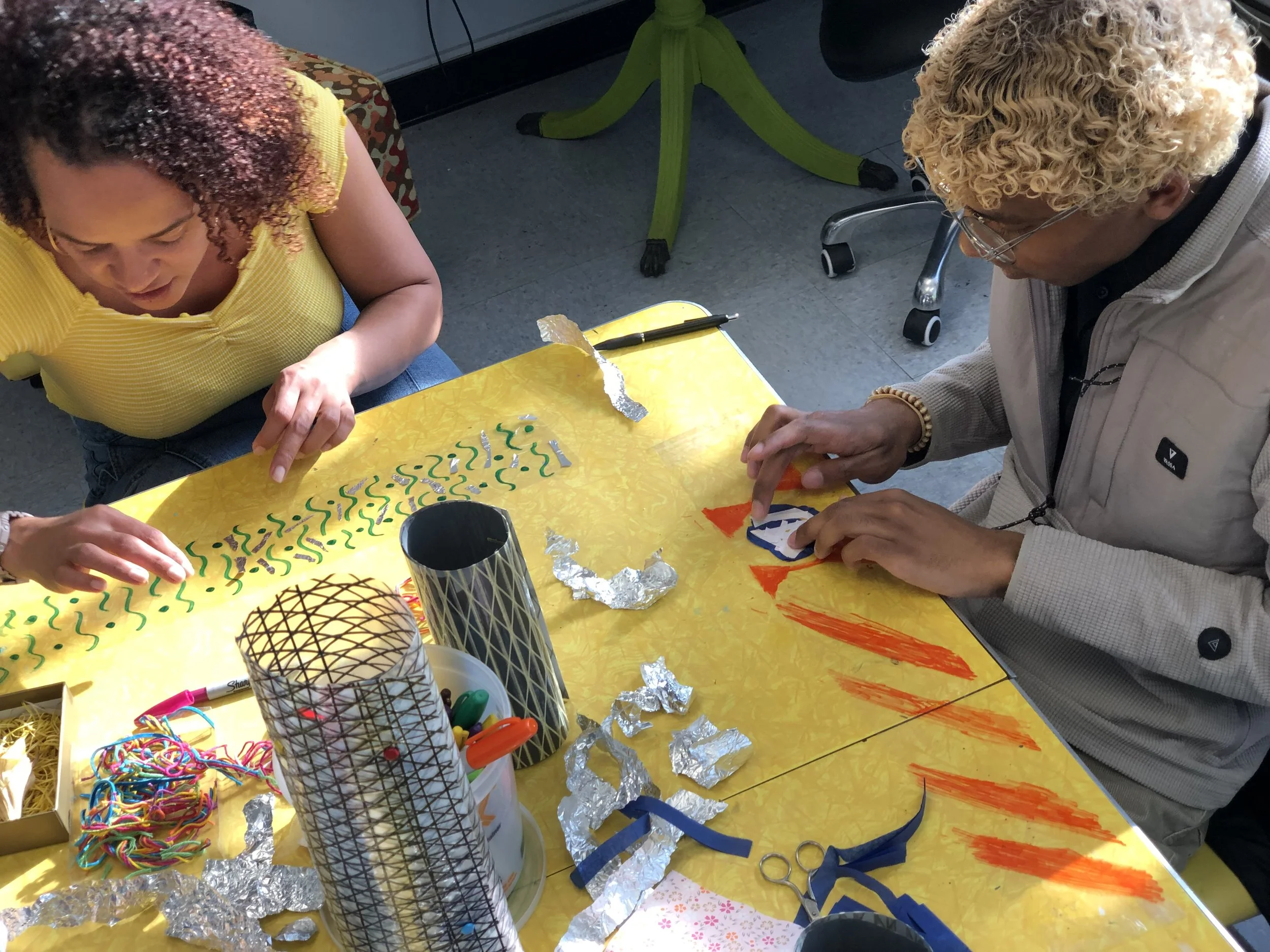 Two people working on a colorful arts and crafts project at a yellow table, surrounded by craft supplies, including foil, yarn, scissors, and markers.