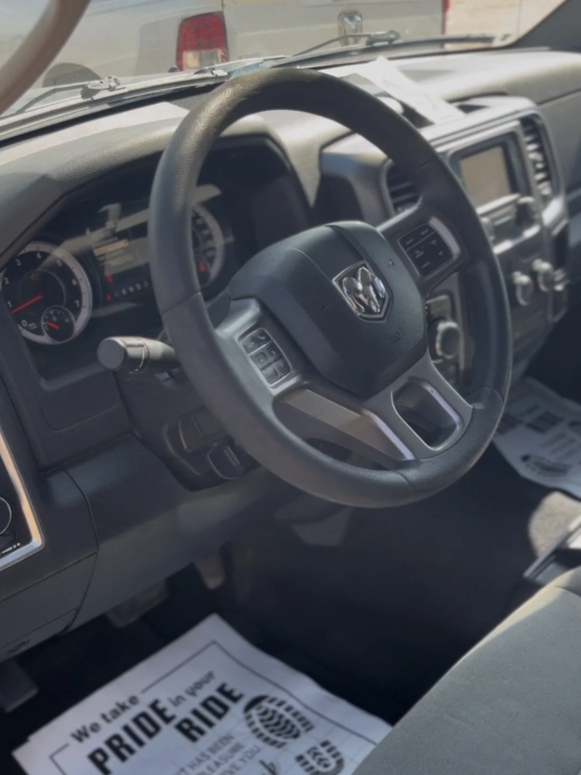 Inside the front of a vehicle showing the steering wheel, dashboard, and instrument panel. There is a paper on the passenger side floor with text about pride and a footprint graphic.