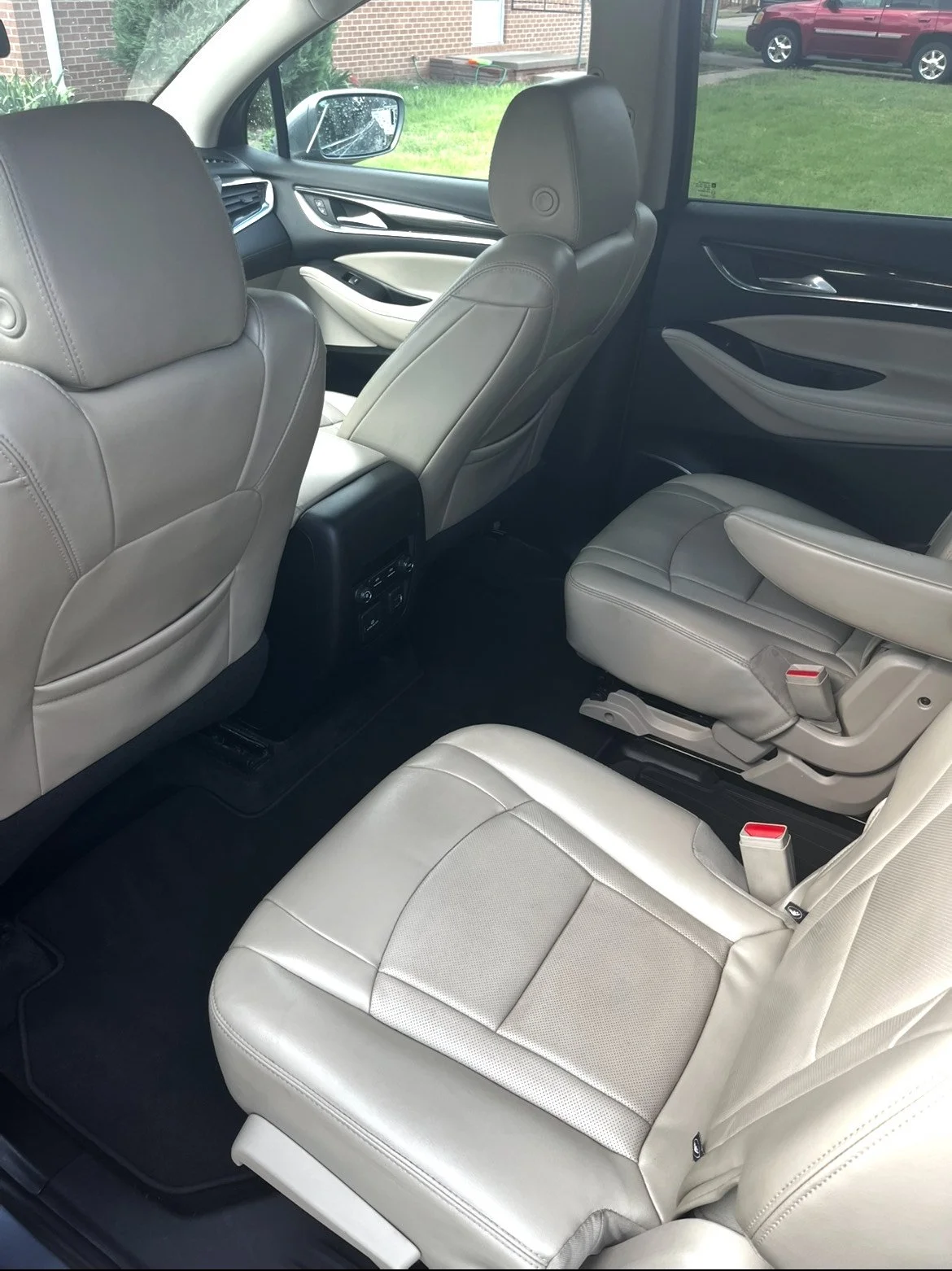 The interior of a vehicle showing beige leather seats and black floor mats, with two rows of seats, including a back row with a beige seat and adjustable armrest and a front row with headrests, door panel, and a window with a grass lawn and a brick b