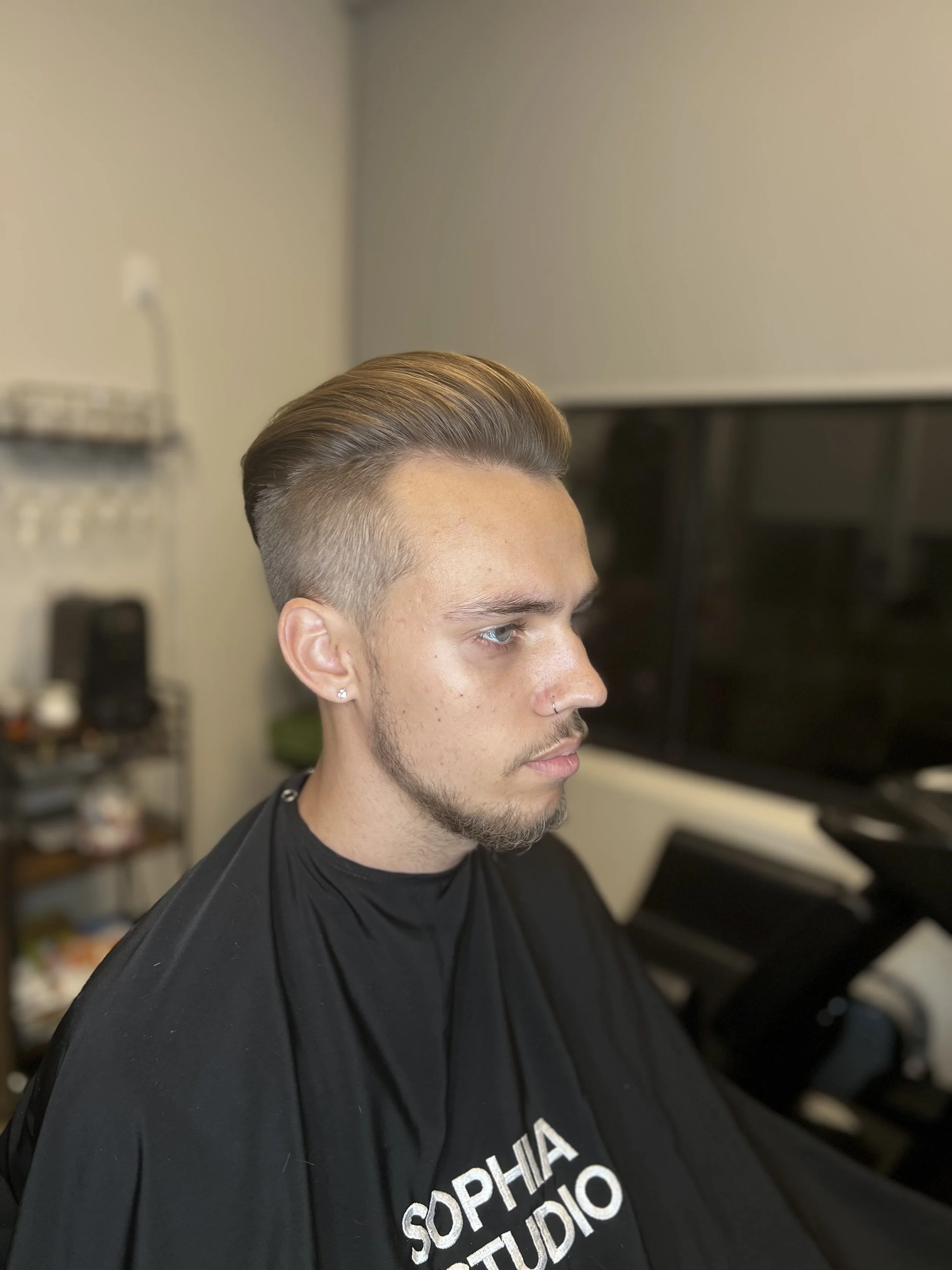 Man with slicked-back hairstyle sitting in a salon chair wearing a black cape with "Sophia Studio" embroidered on it.