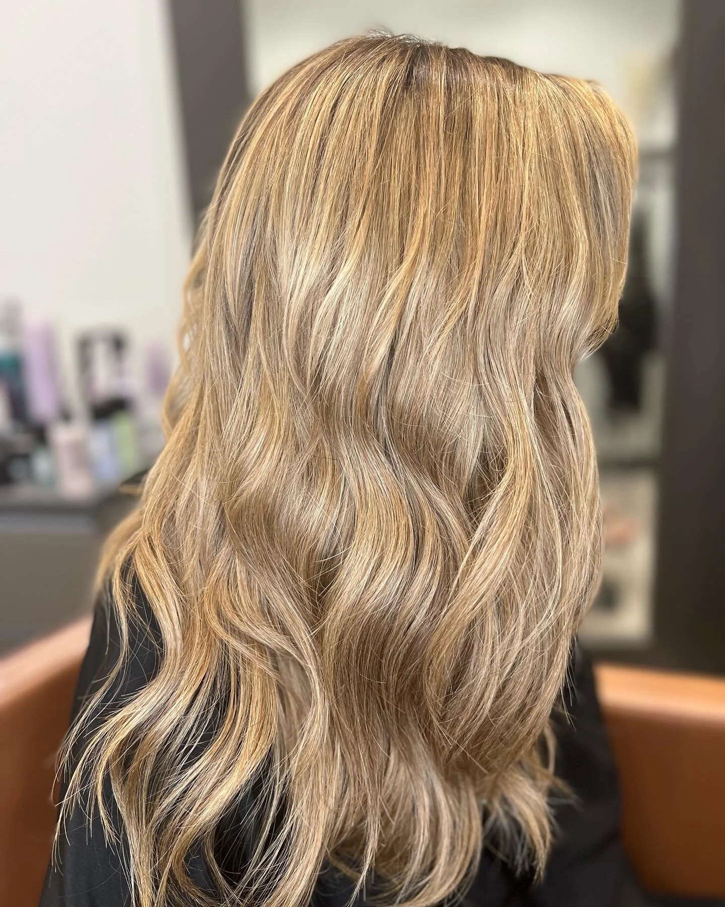 Close-up of wavy blonde hair cascading down the side and back of a person, showing a salon setting in the background.