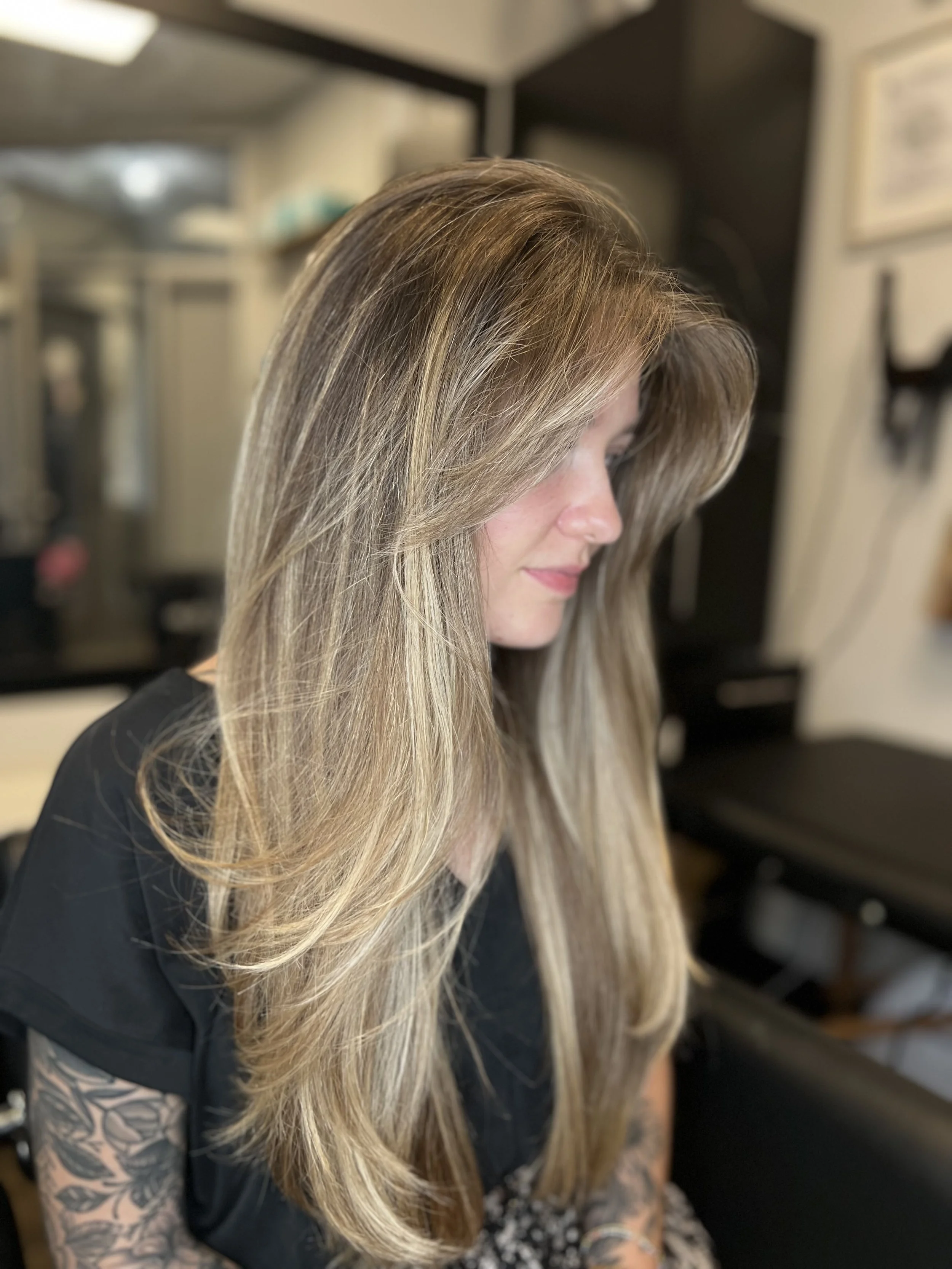 Woman with long, layered blonde hair sitting in a salon chair, tattooed arm visible, blurred salon background.