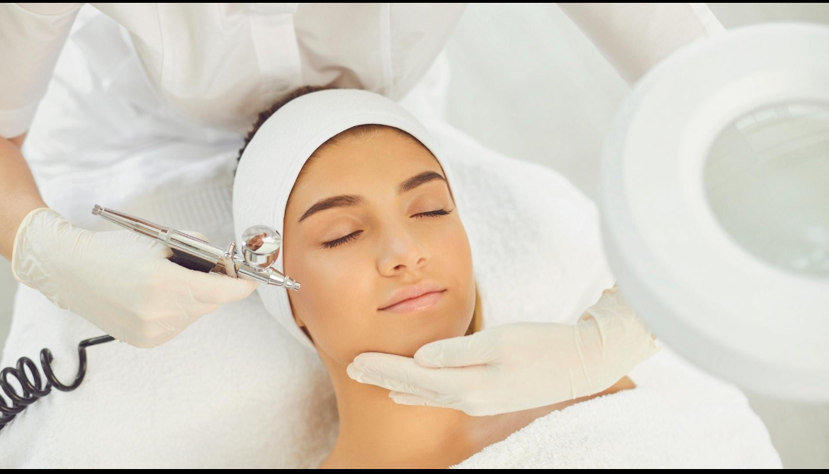 Woman receiving a facial airbrush treatment by a beautician wearing gloves in a spa setting.