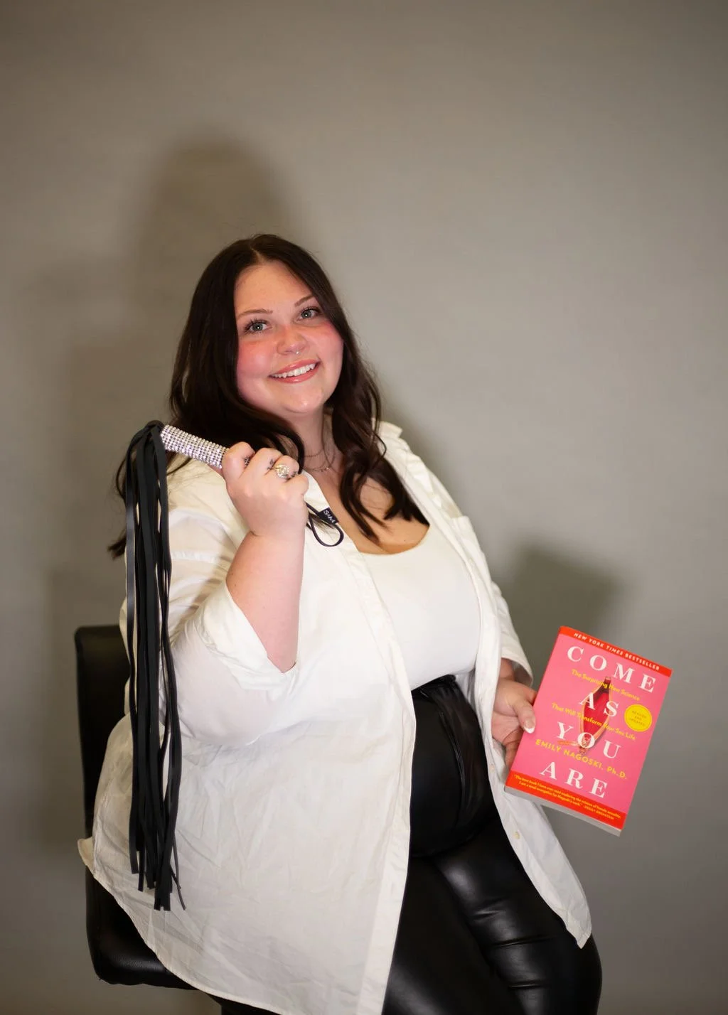 A smiling woman with dark hair and a white shirt holding a book titled "Come As You Are" by Emily Nagoski in a room with plain wall.