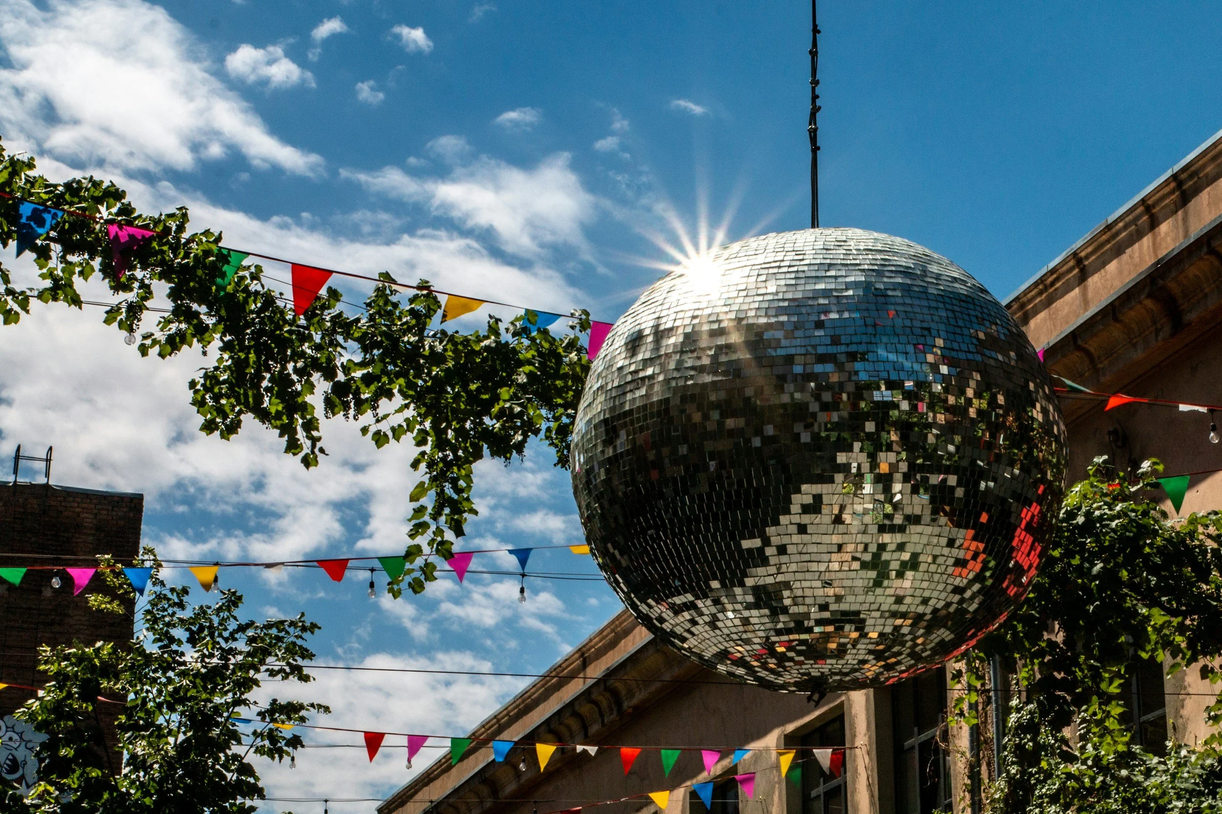 A large mirror disco ball hanging outdoors with strings of colorful pennant banners, tree branches, a blue sky with some clouds, and part of a building.