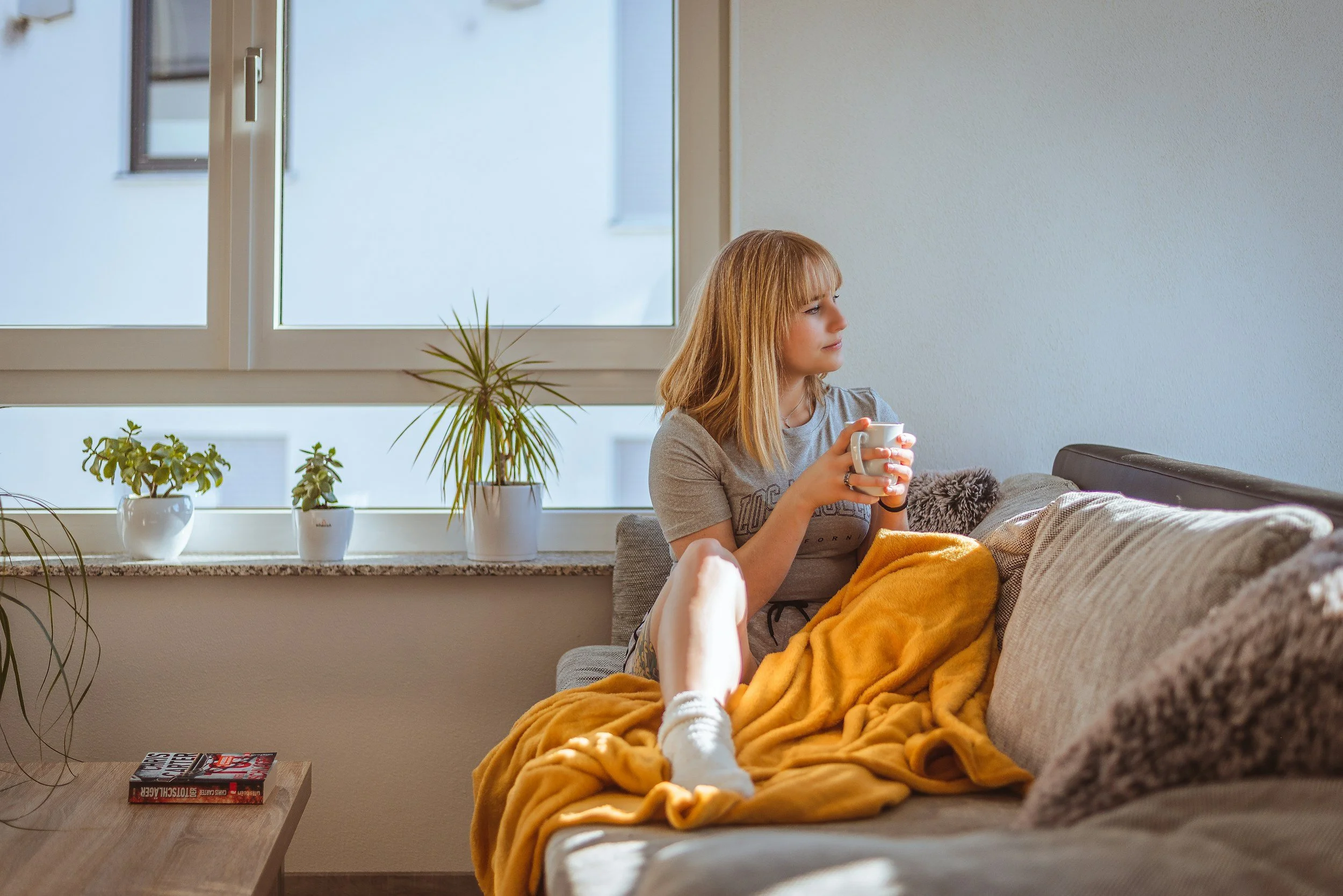 A young woman sitting on a beige sofa with a yellow blanket, holding a mug and looking out the window in a bright living room with potted plants on the windowsill and a book on the wooden side table.