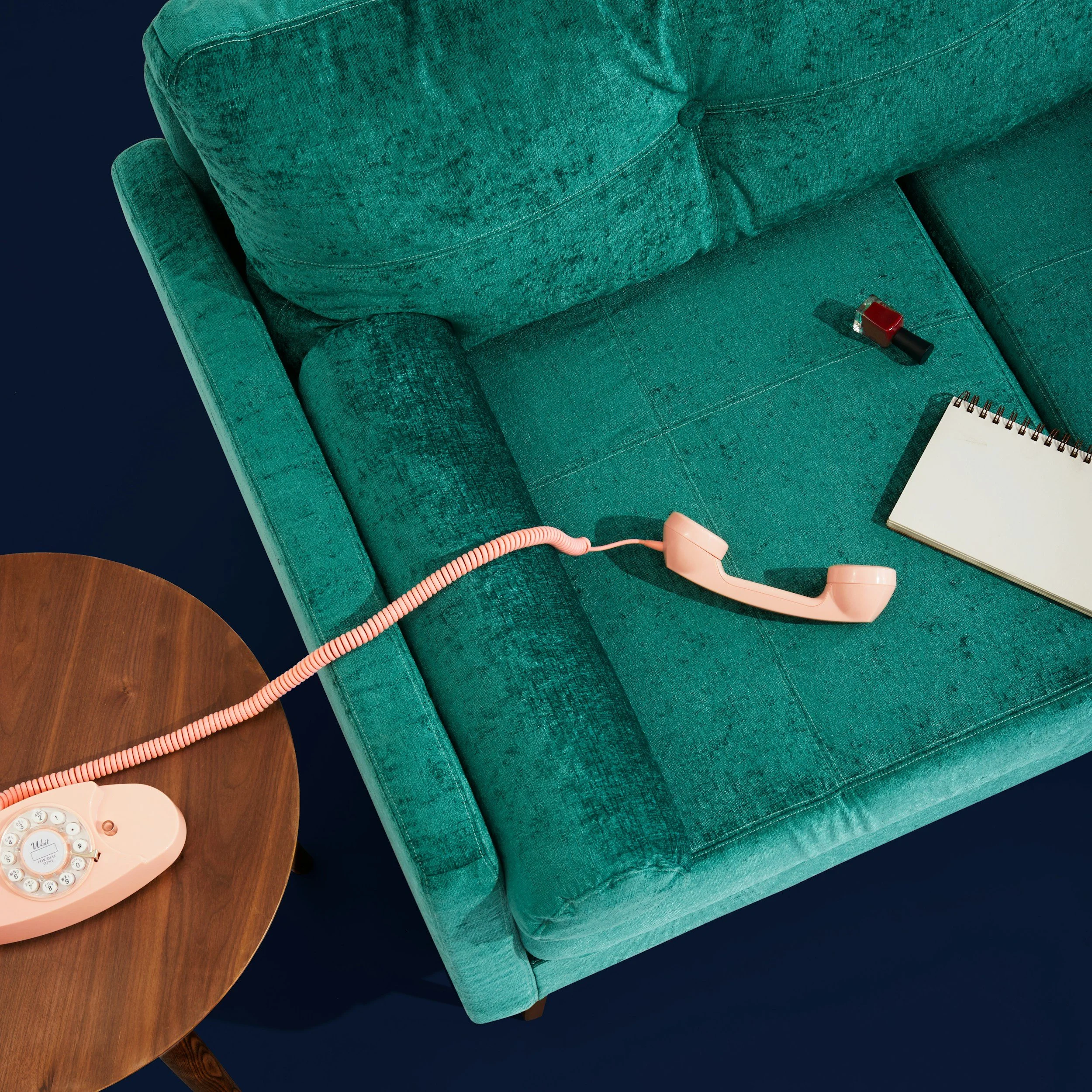 A green vintage sofa with a pink rotary phone, a small notepad, and a lipstick placed on it, beside a round wooden table.