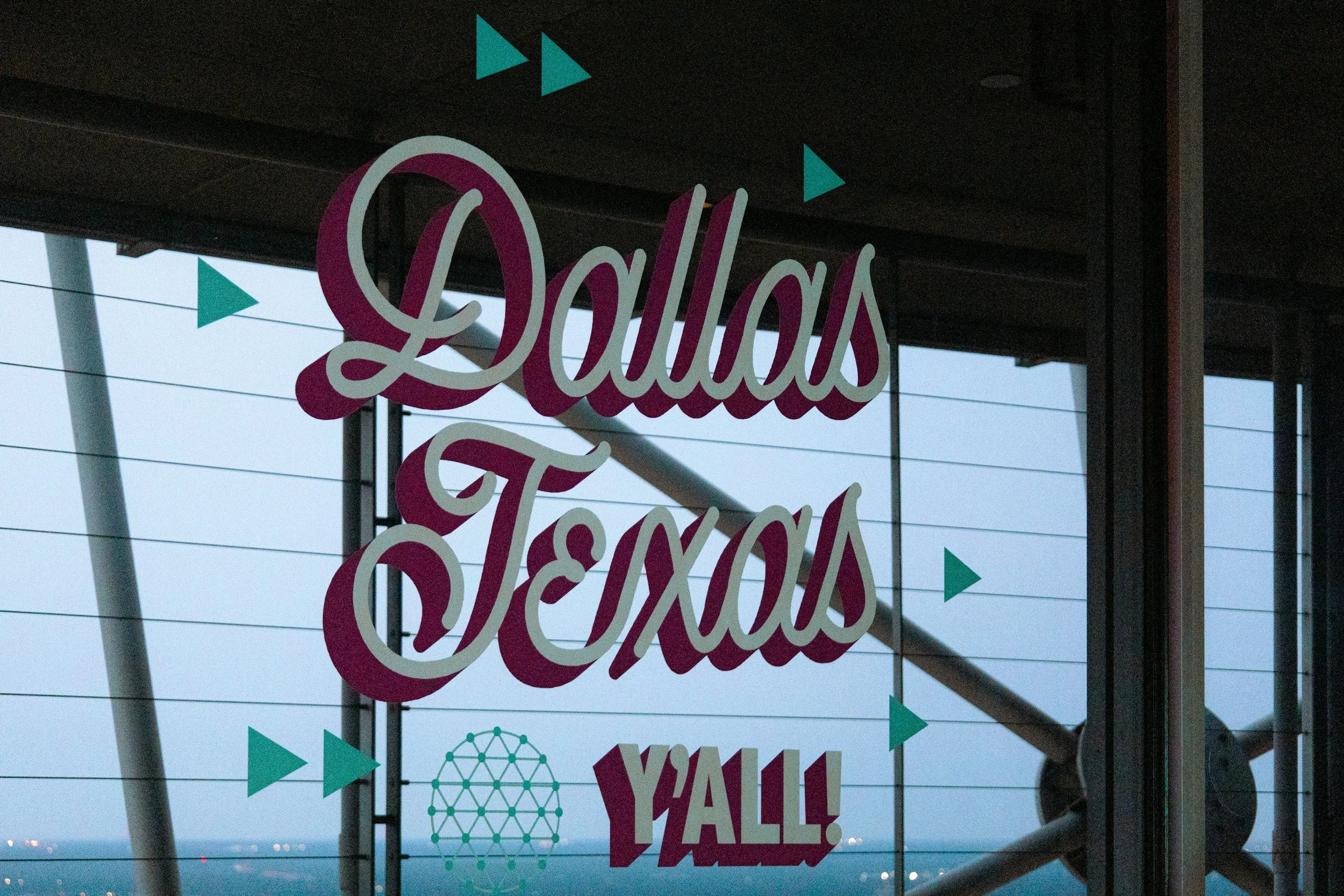 A window with a colorful sign that says 'Dallas Texas Y'all' with small teal triangles and a teal globe pattern in the background, viewed from inside a building with metal beams.