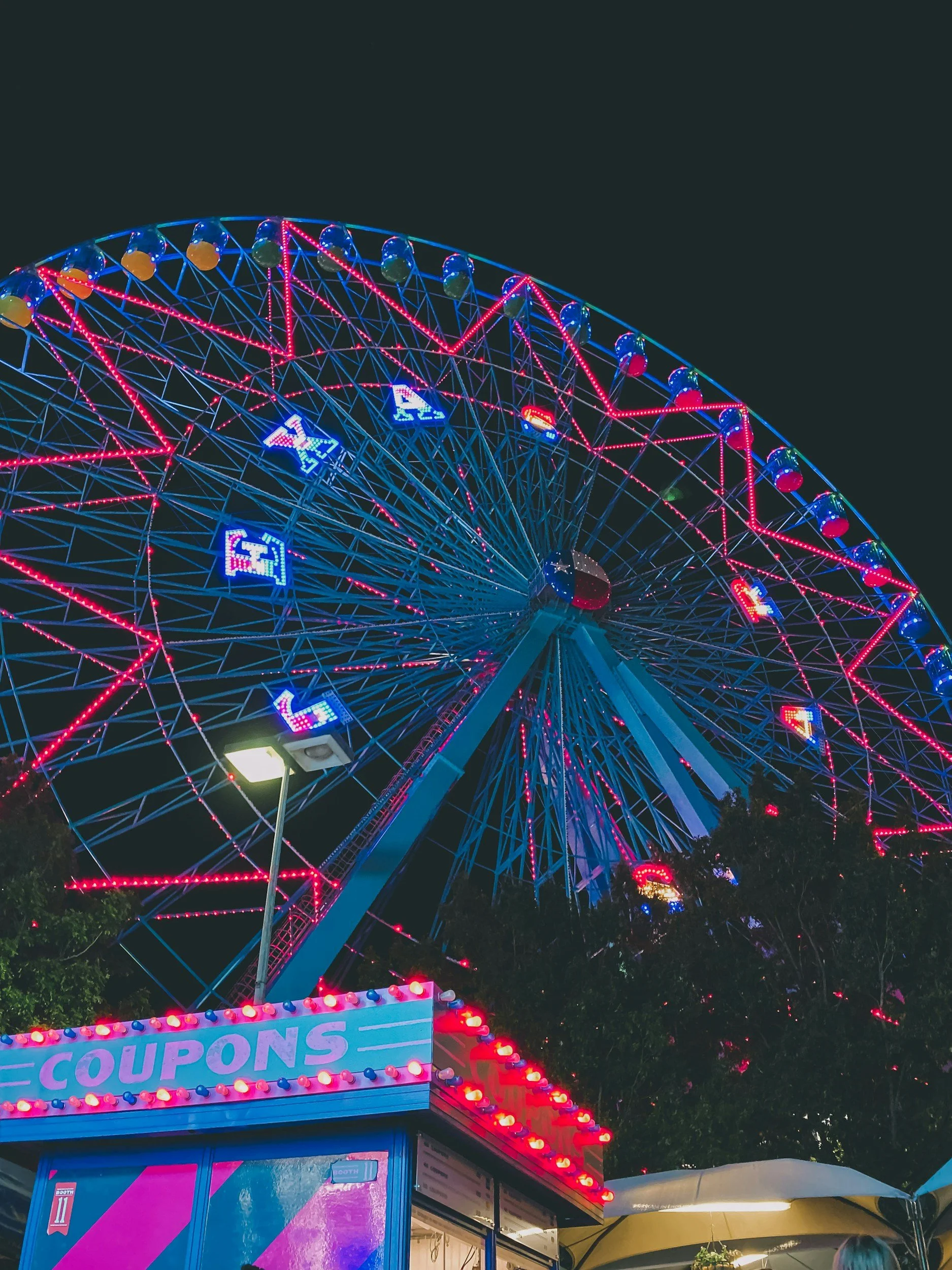 Colorful illuminated ferris wheel at night near a booth with a sign that says "COUPONS".