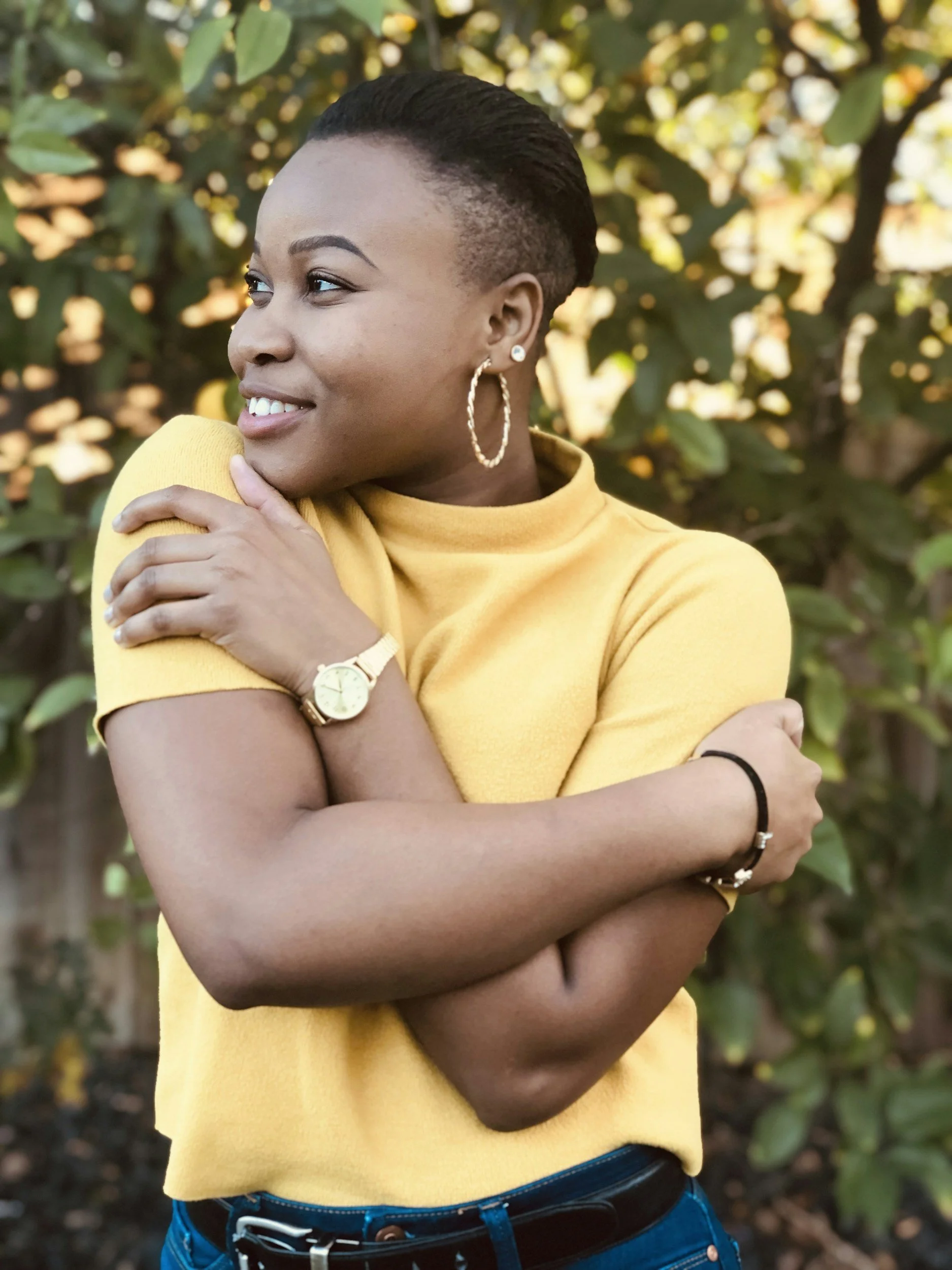 A woman with short hair, wearing a yellow top, gold watch, hoop earrings, and a black bracelet, smiling and hugging herself in front of greenery with sunlight filtering through the leaves.