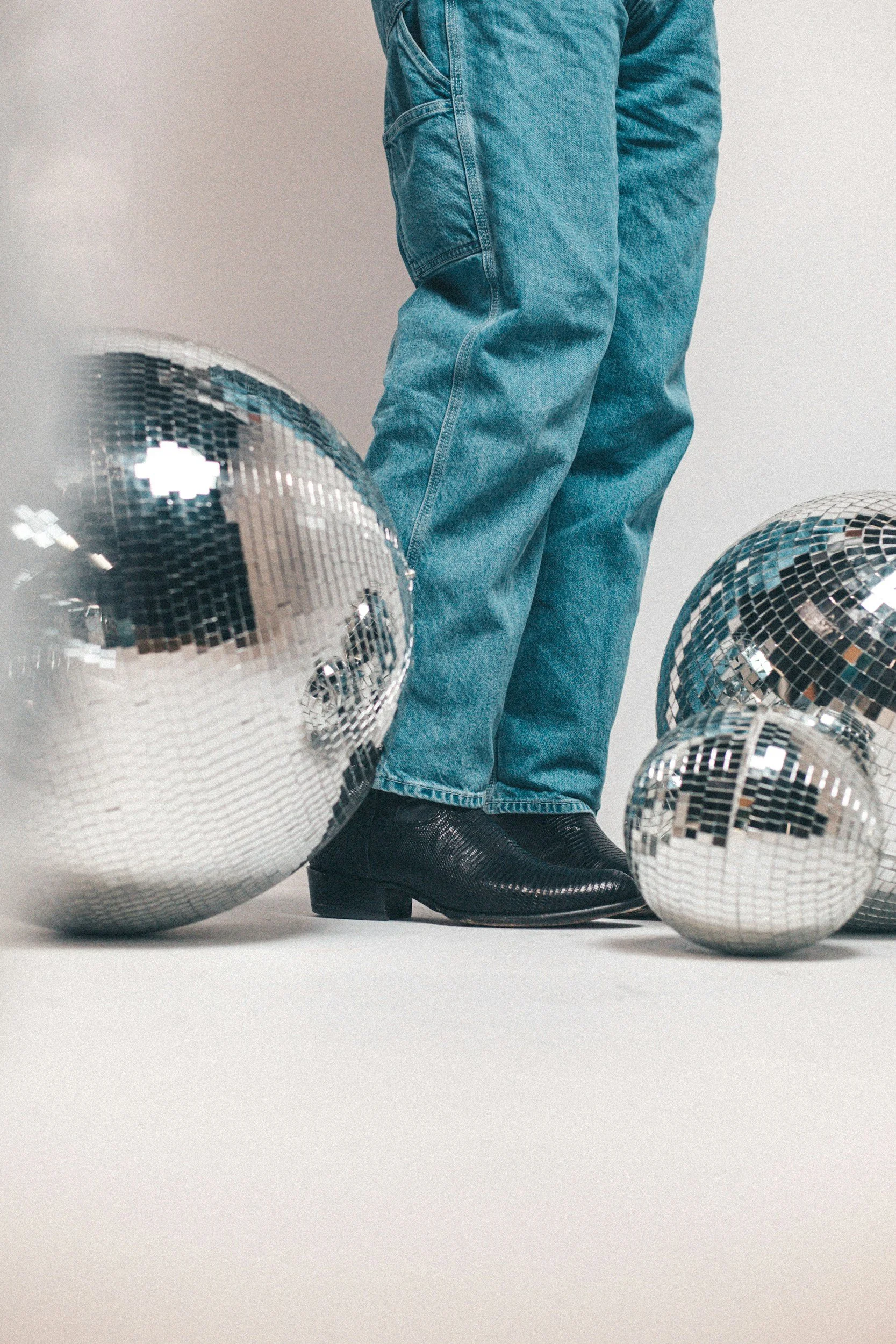 Person standing among disco balls, wearing blue jeans and black boots.