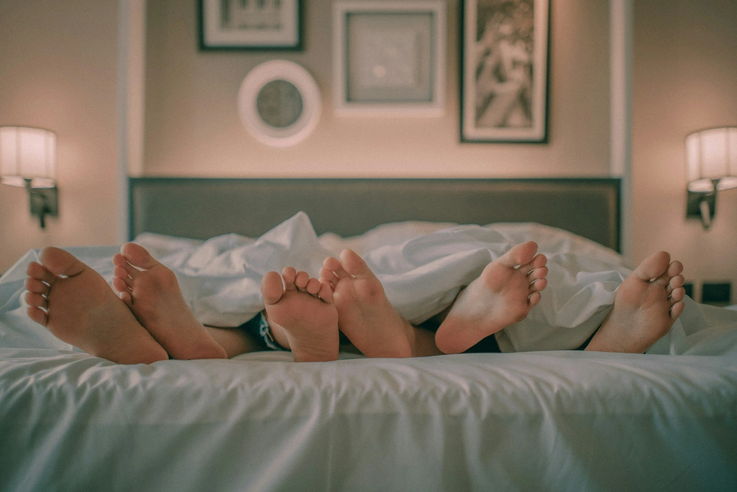 People lying in bed with their feet facing the camera, with framed pictures on the wall and two lamps on either side of the bed.