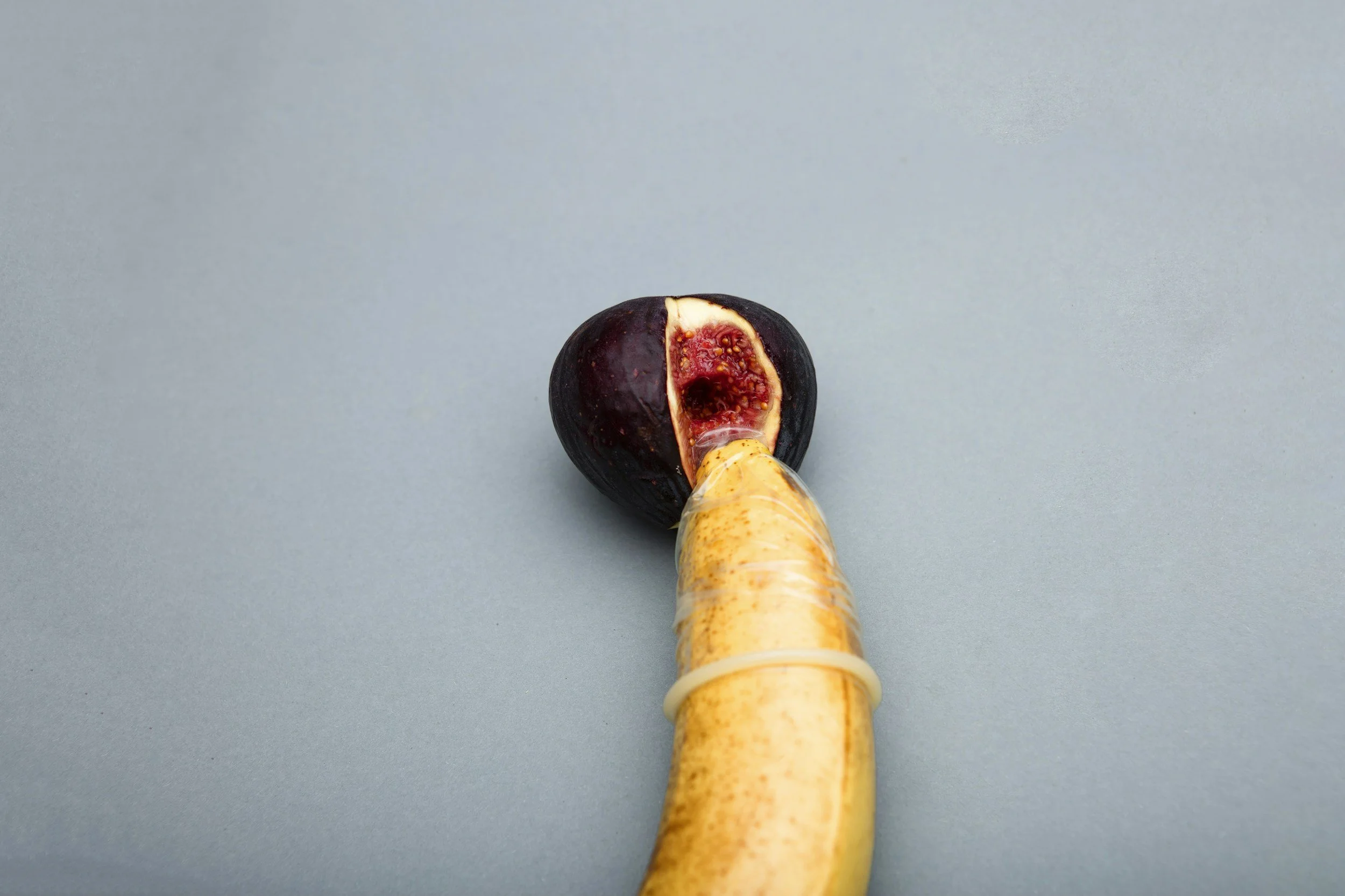 A ripe purple fig with a cut revealing red flesh and yellow seeds, on top of a yellow elongated stalk, against a plain light blue background.