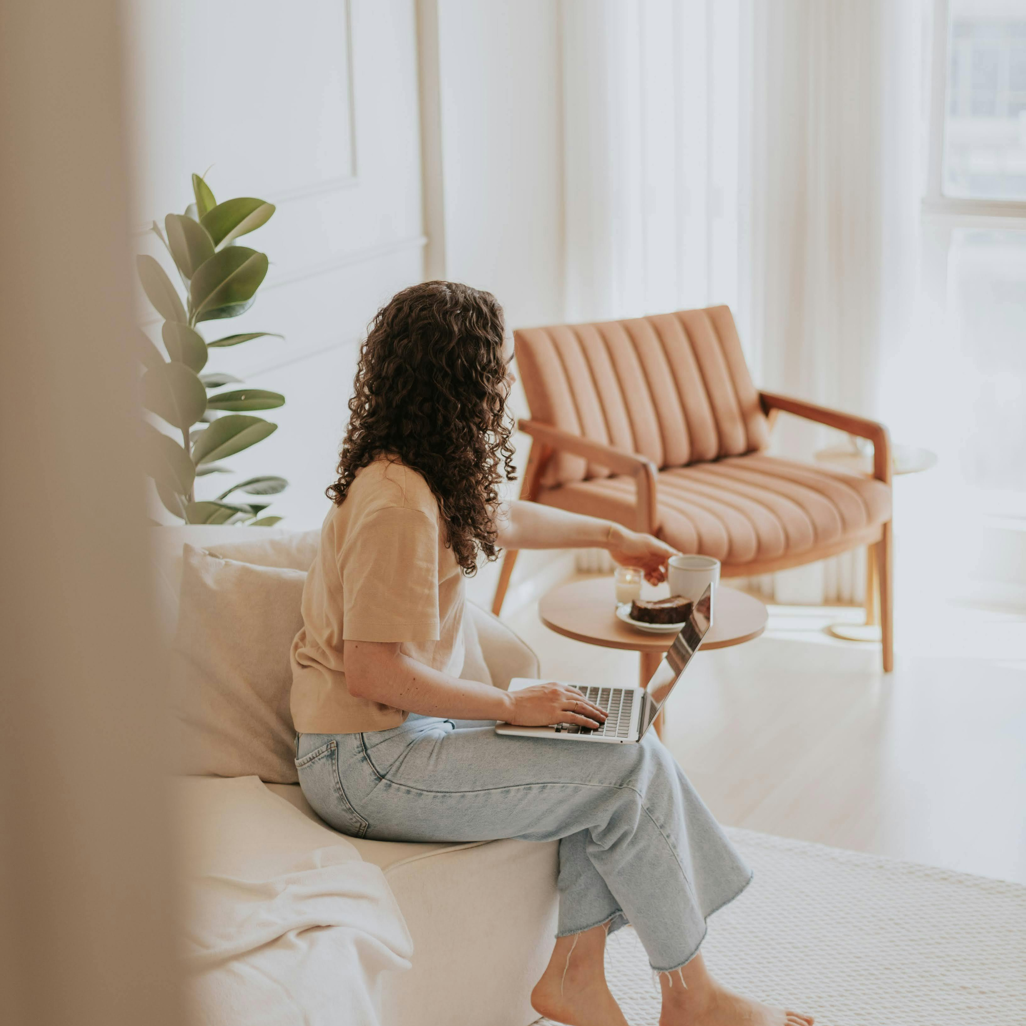 A woman with curly hair sitting on a beige sofa, working on a laptop with a coffee mug and cake nearby, in a bright, cozy living room with a peach-colored armchair and a large window.