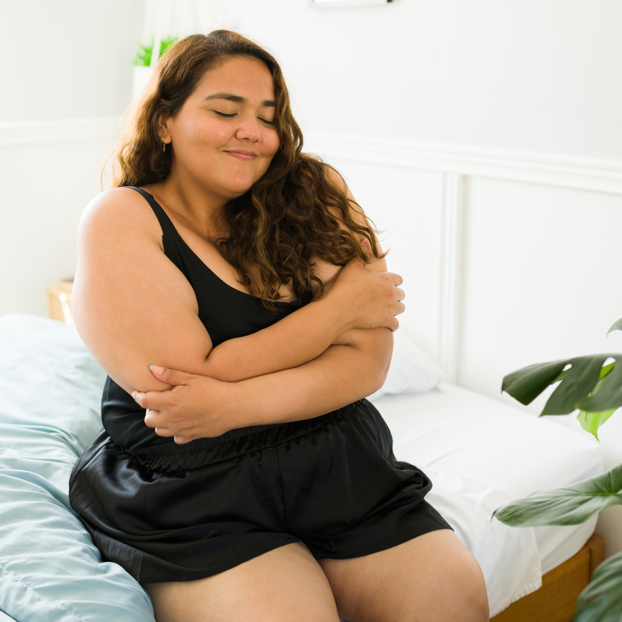 A woman sitting on a bed, hugging herself with her eyes closed and smiling peacefully.