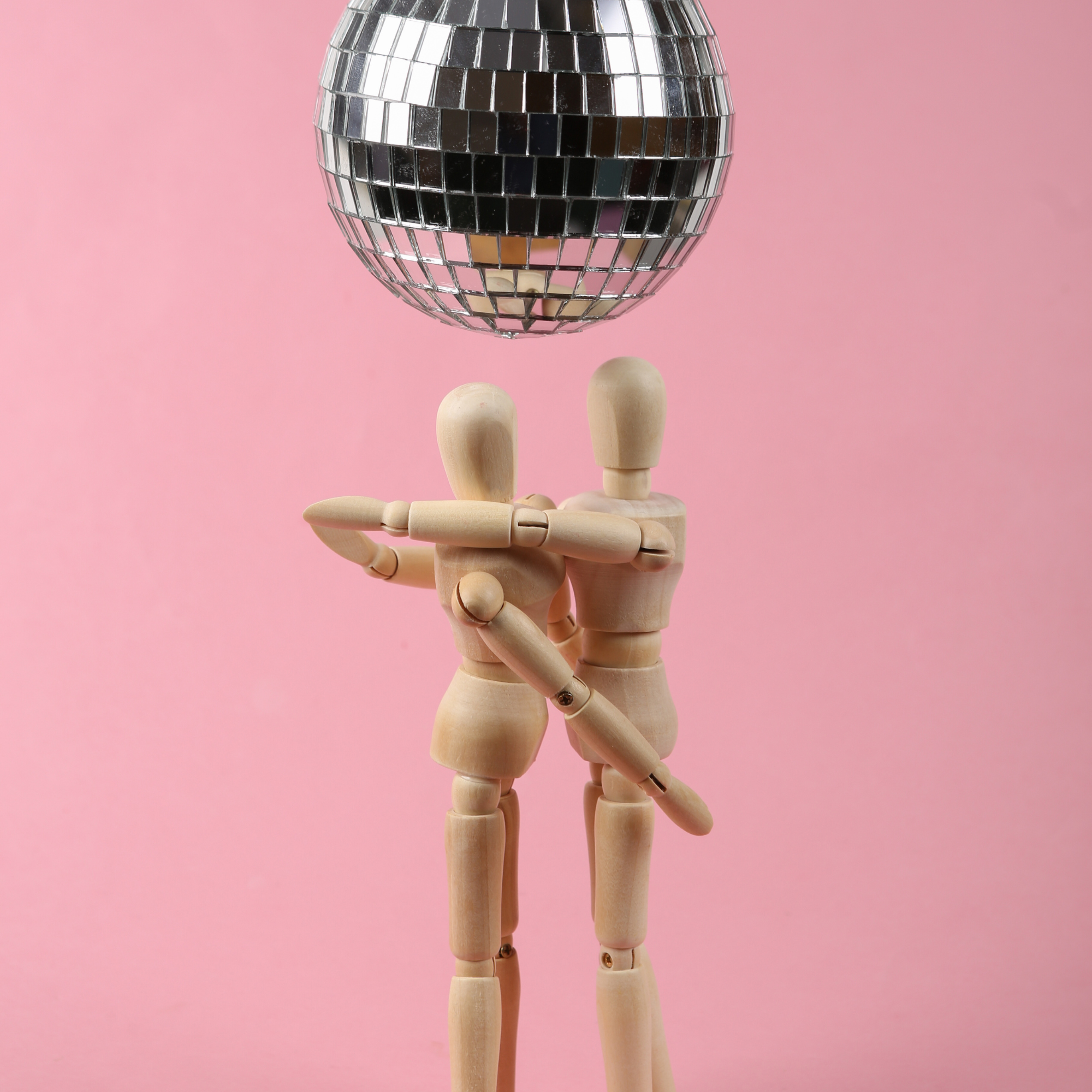 Two wooden artist mannequins dancing under a disco ball against a pink background.