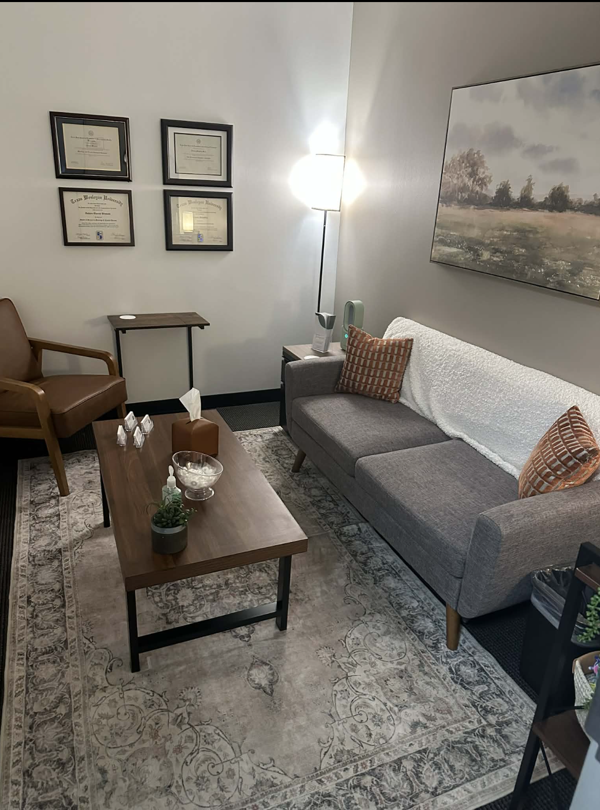A cozy waiting room with a gray sofa with two orange patterned pillows, a wooden coffee table with a potted plant, tissue box, sanitizer, and a glass bowl, a brown armchair, a small side table with a lamp, artwork on the wall, and framed certificates, all illuminated by a floor lamp.