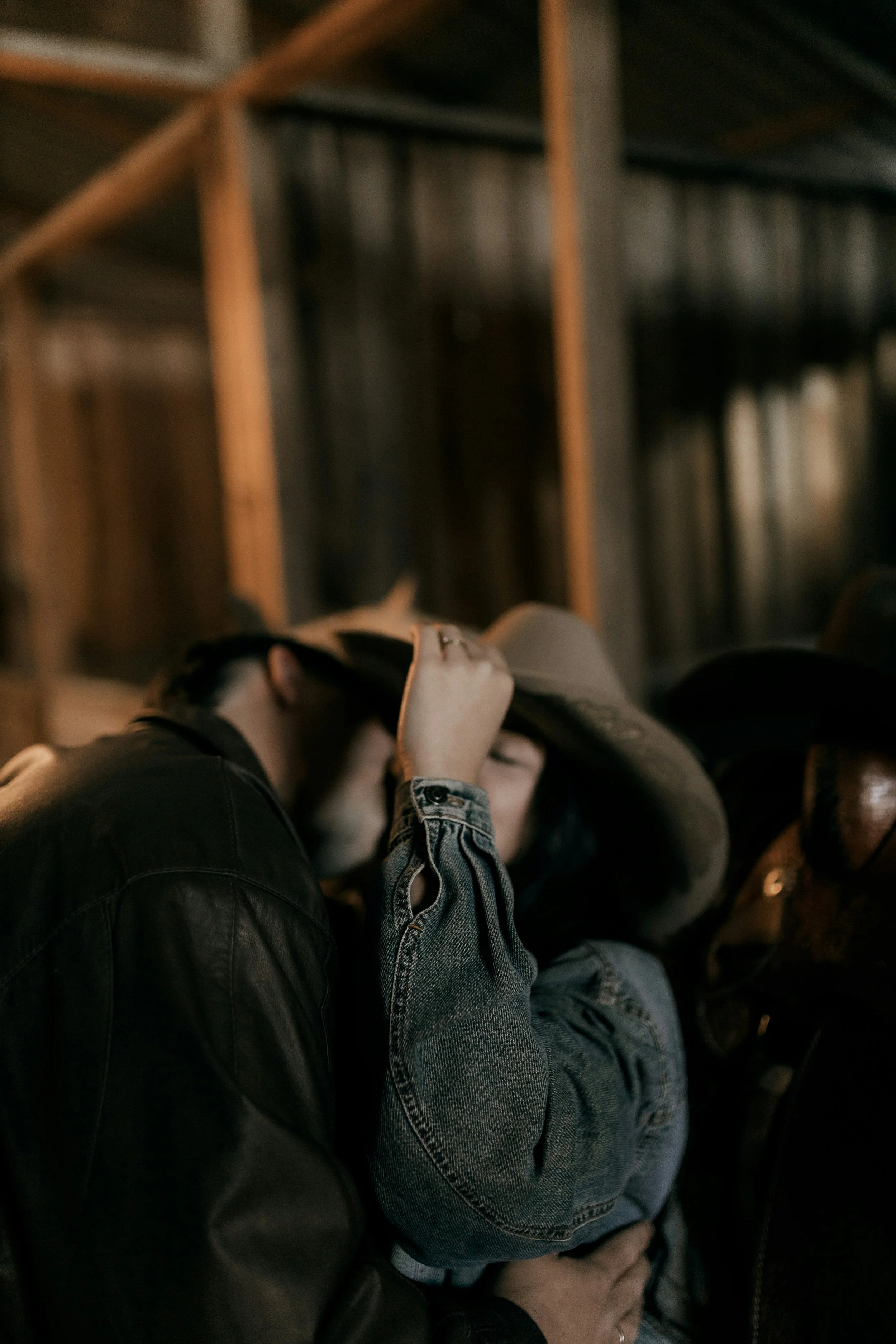 A couple sharing an intimate moment, with the woman wearing a large hat and denim jacket, in a dimly lit indoor setting.