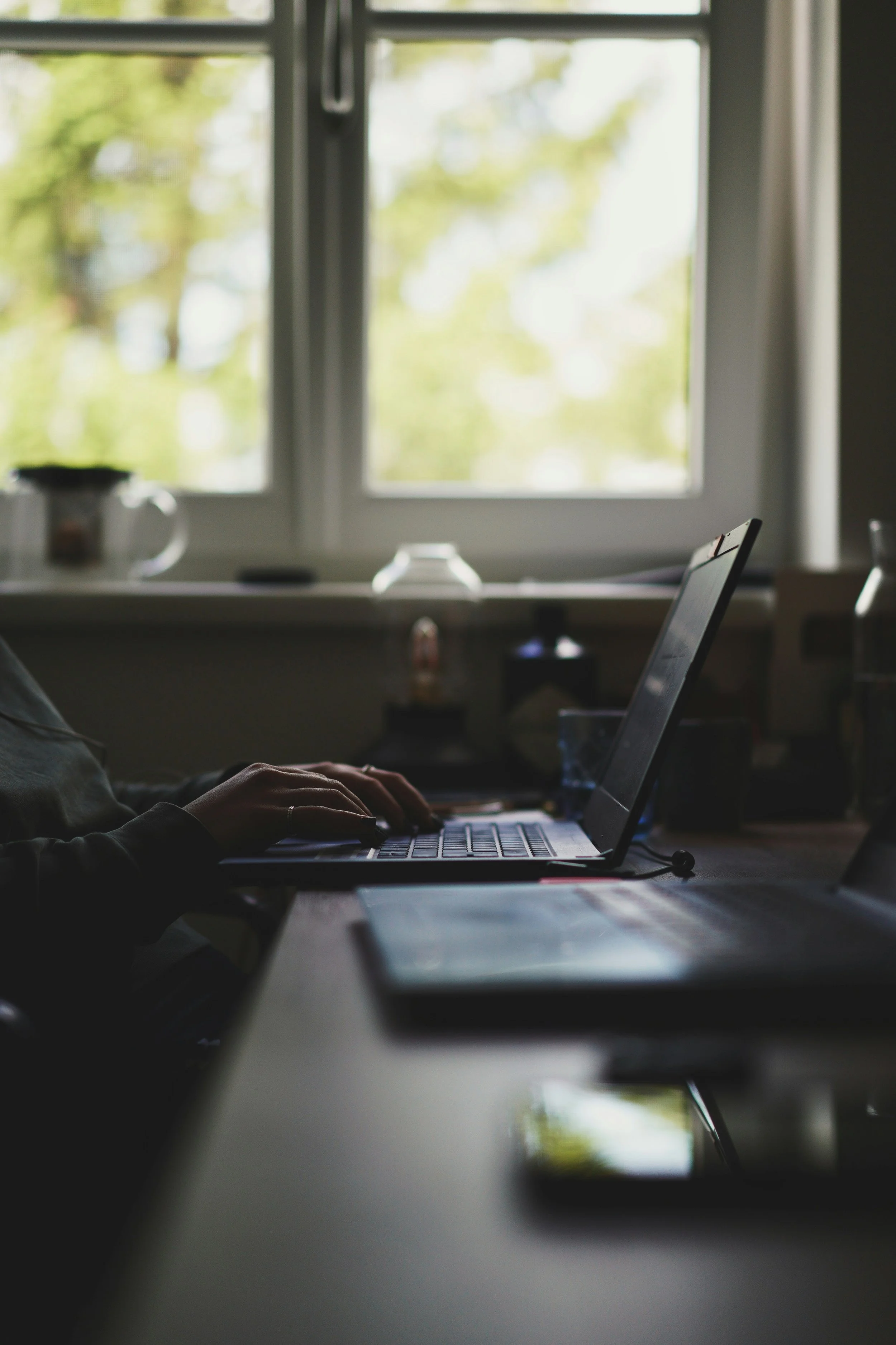 Person using a laptop at a desk near a window with green trees visible outside.