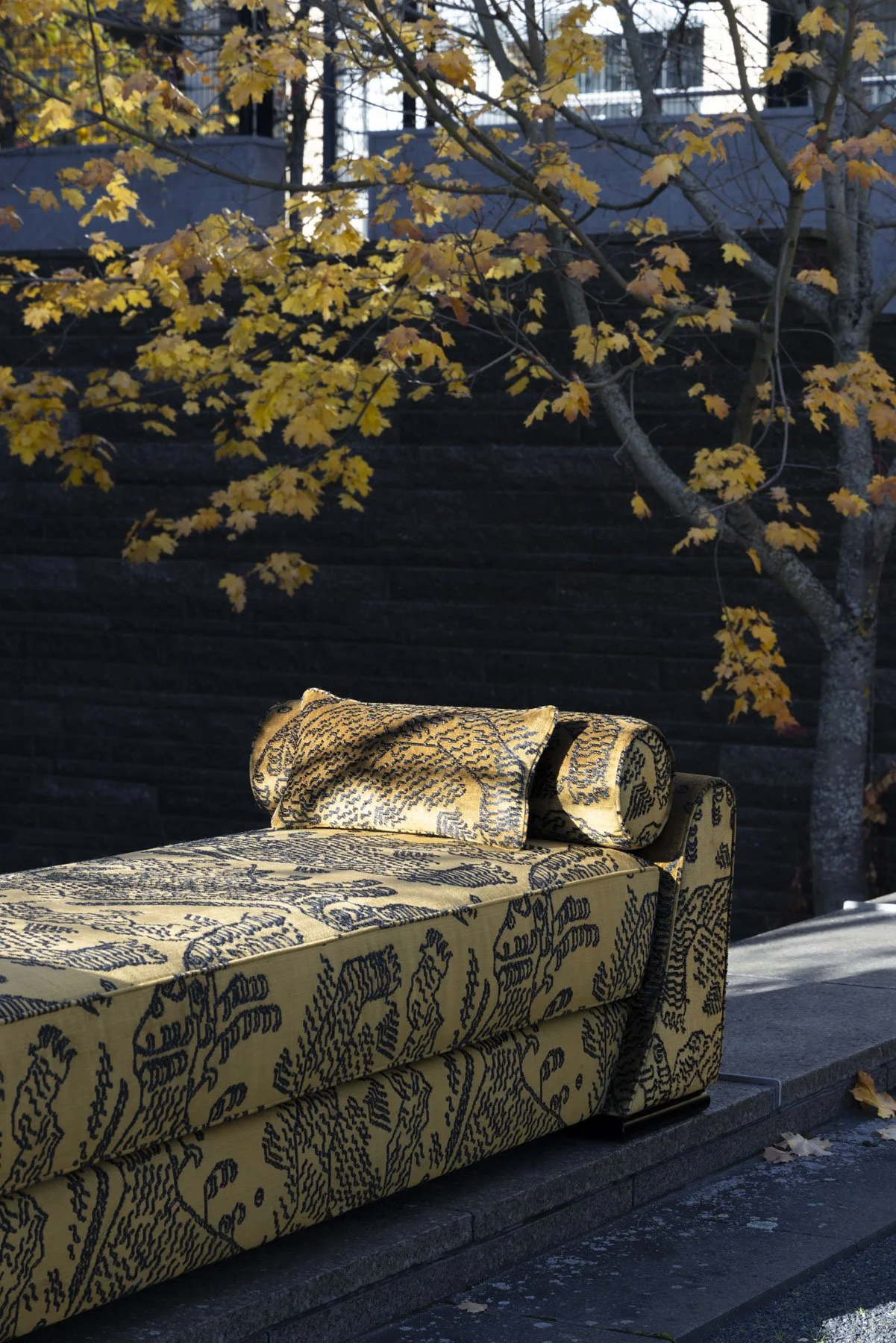 Yellow and black patterned Borghese longue with a matching pillow, designed by Carola Rytsölä, set outdoors on a stone surface near a tree with yellow leaves and a dark wall in the background. Tiger fabric by Dedar Milano.