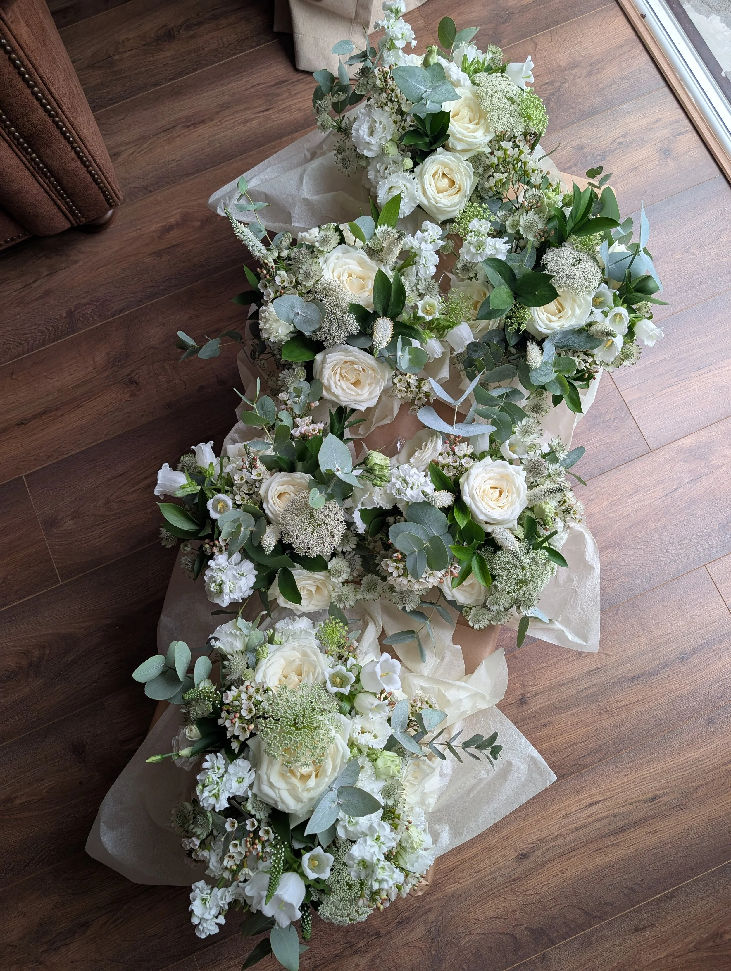 Multiple white floral arrangements with roses, greenery, and other white flowers on a wooden floor near a window.