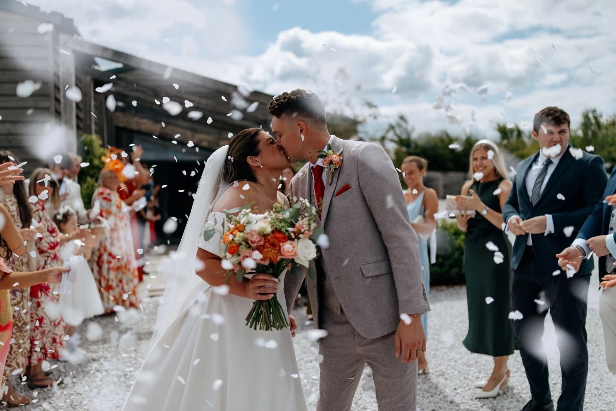 A newlywed couple shares a kiss outside during a wedding celebration, surrounded by guests throwing flower petals.