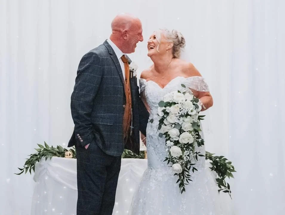 An elderly couple, dressed in wedding attire, smiling and looking at each other affectionately during their wedding ceremony. The woman is holding a large bouquet of white roses and greenery.