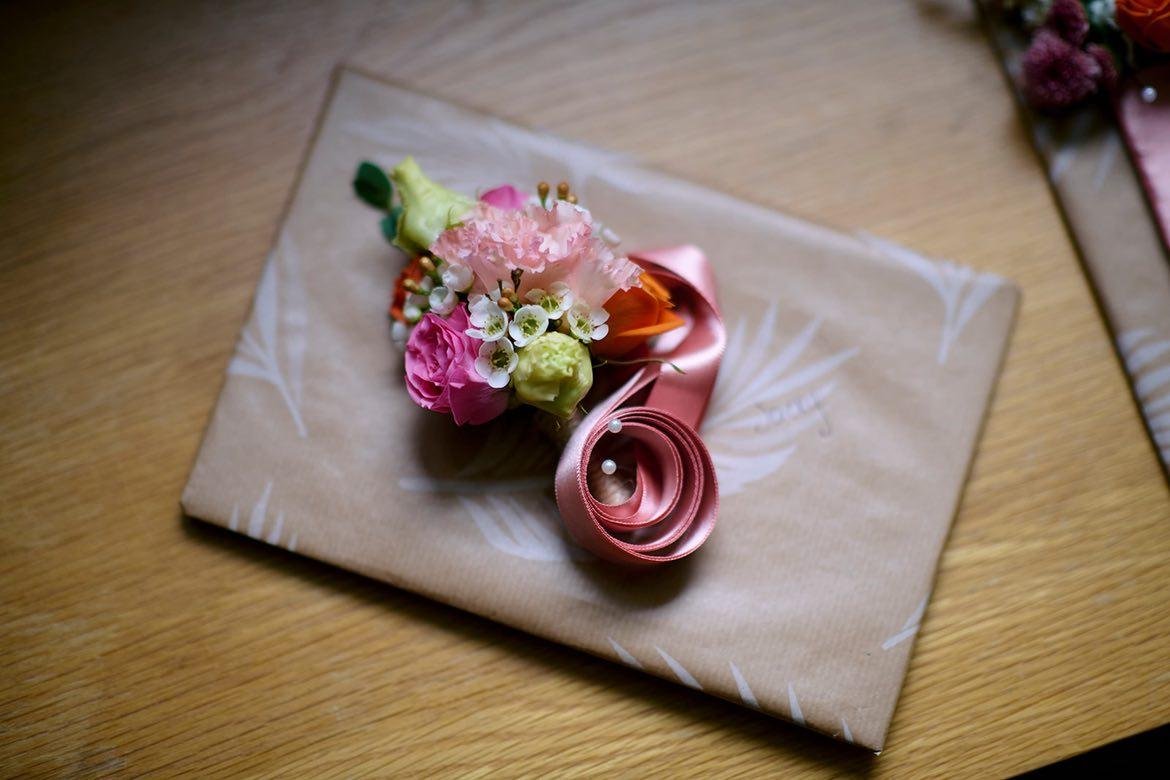 A boutonniere with pink, peach, and white flowers, wrapped with pink satin ribbon, resting on a beige gift box with white feather patterns on a wooden surface.