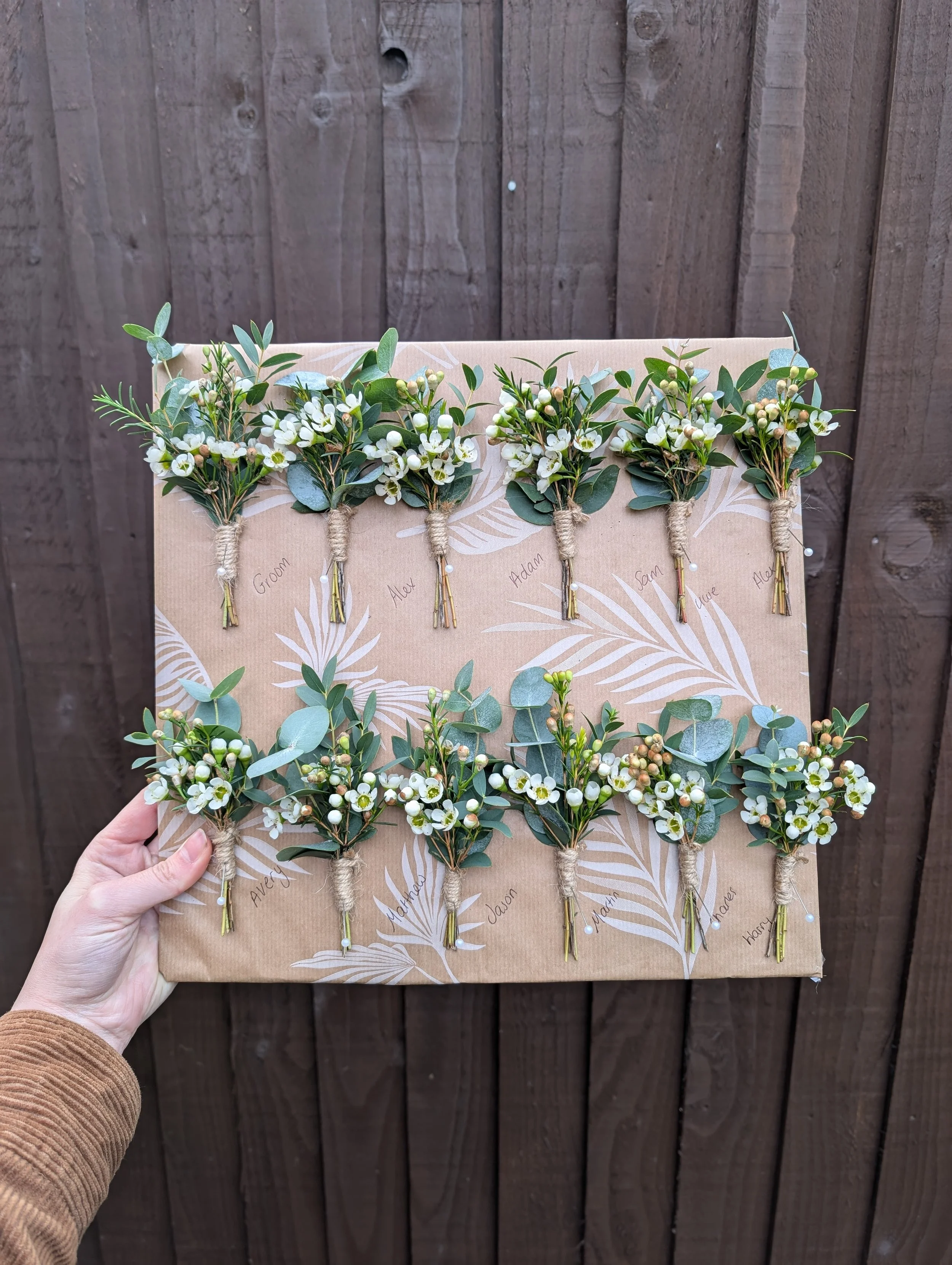 Flat lay of twelve small boutonniere floral arrangements with white flowers and greenery, labeled with handwritten names, arranged on a cardboard backing against a wooden fence.