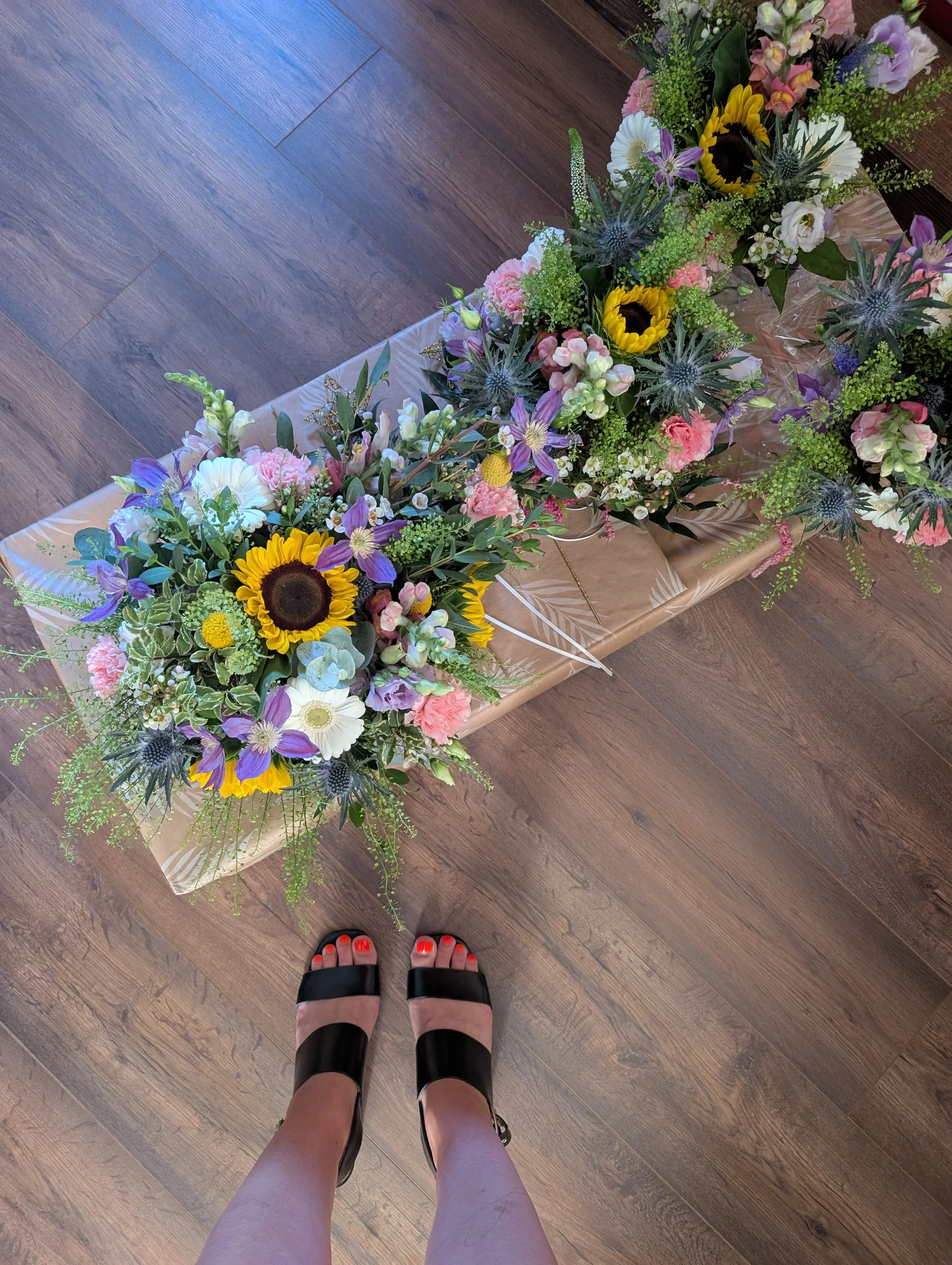 View of two floral arrangements with sunflowers, purple, pink, and white flowers, and greenery on a wrapped table, seen from the perspective of someone standing in black and pink sandals on wood flooring.