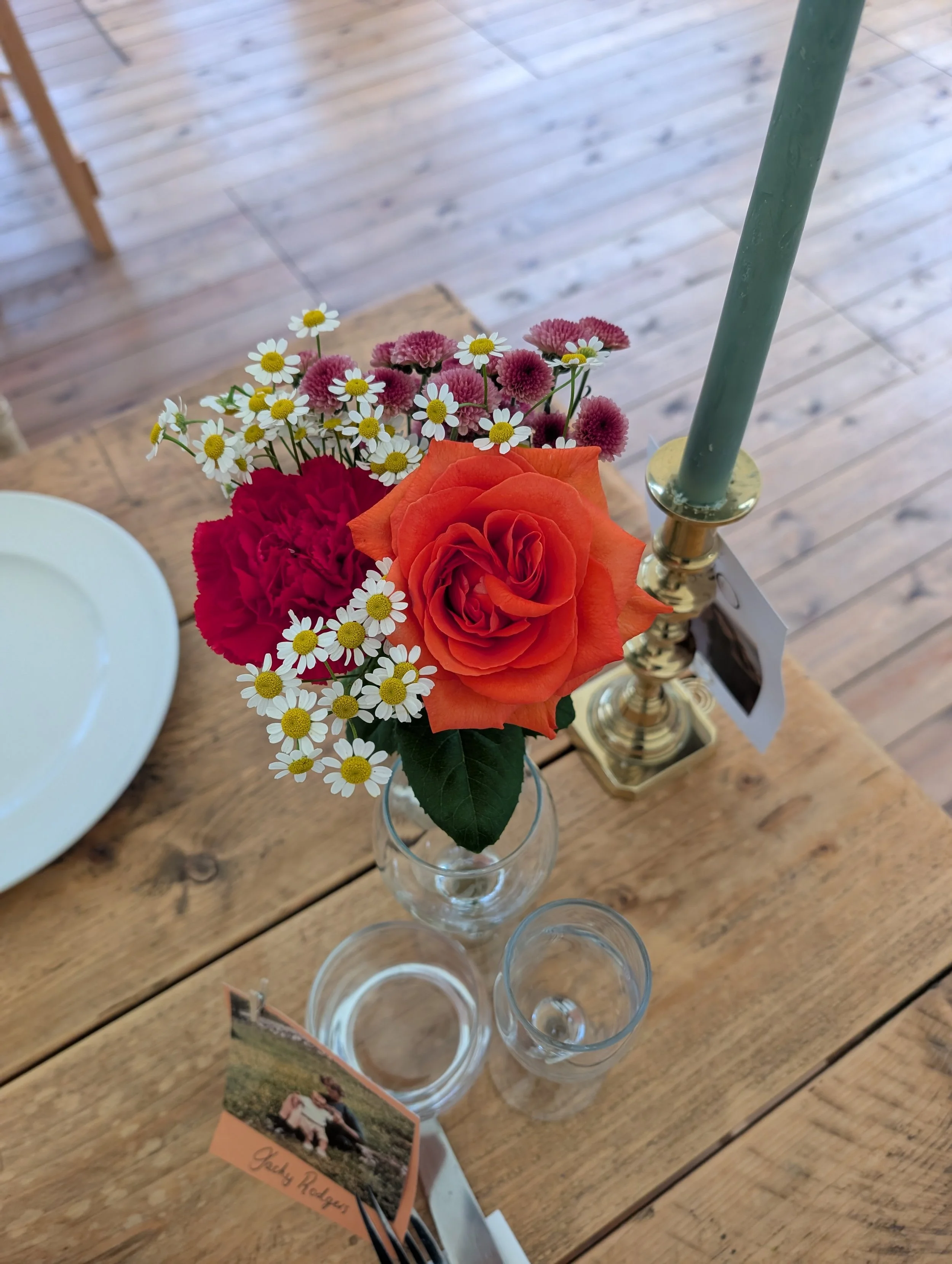 A small flower arrangement with a large orange rose, red carnations, and small white daisies with yellow centers in a glass vase. Next to it is a brass candle holder with a green candle. The flowers are on a wooden dining table with a white plate and a name tag.