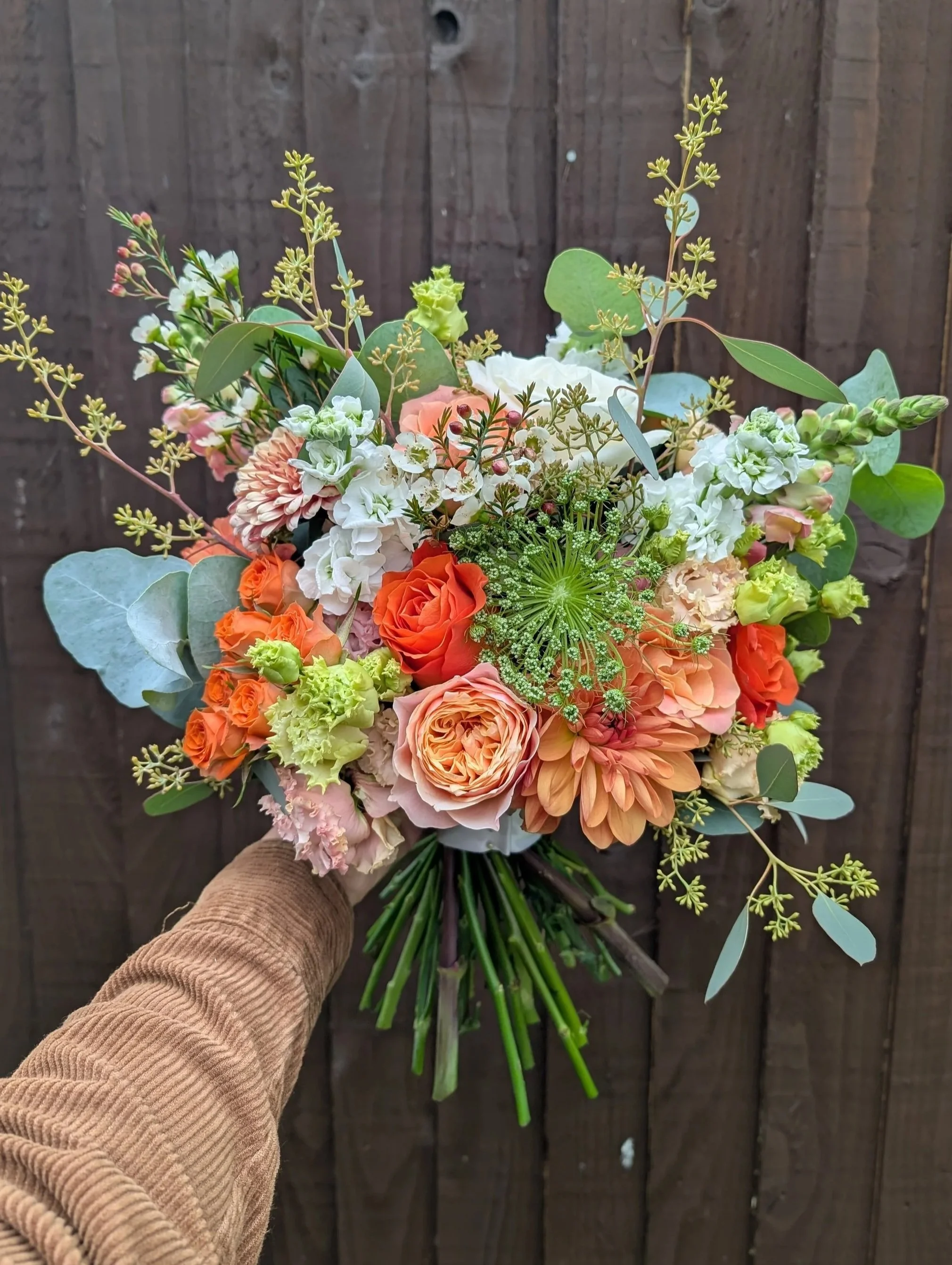 Hand holding a bouquet of mixed flowers including roses, dahlias, and greenery against a wooden background.