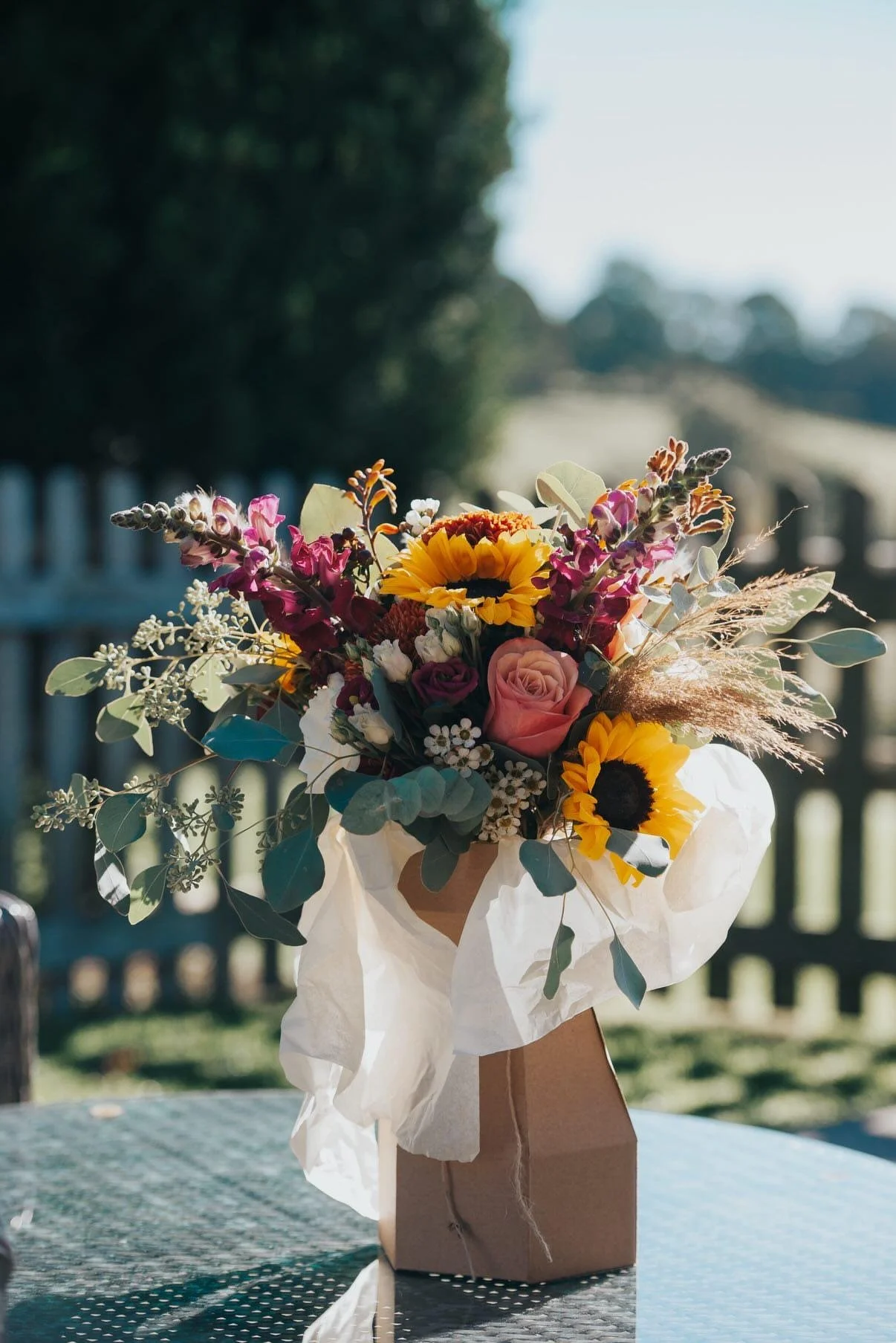 A bouquet of mixed flowers including sunflowers, roses, and other wildflowers in a brown paper bag on a table outdoors.