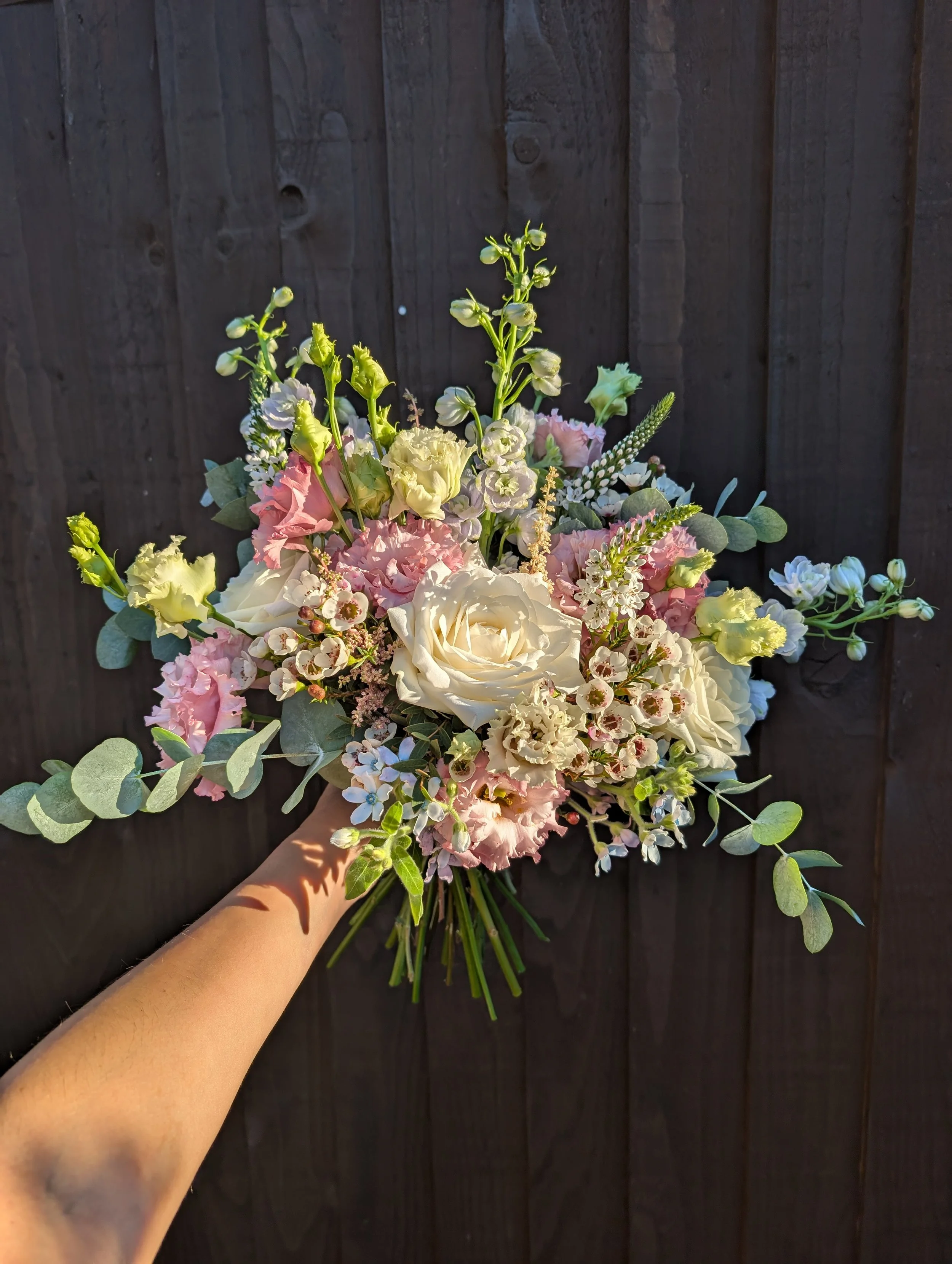 Person holding a bouquet of pink, white, and green flowers in front of a wooden fence.