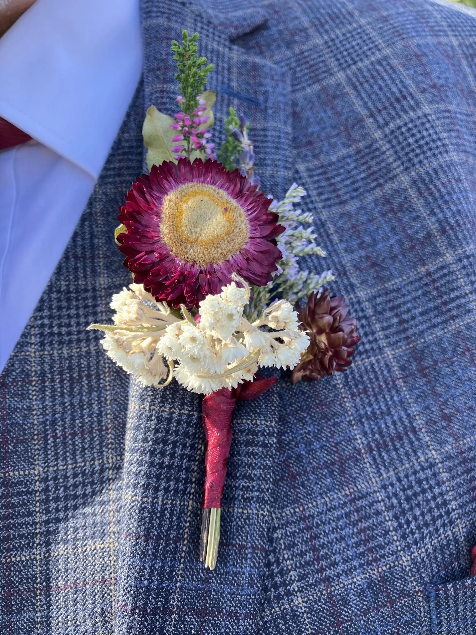 A boutonniere with a large purple and yellow flower, white flowers, and greenery, wrapped with red ribbon, placed on a person's blue suit.
