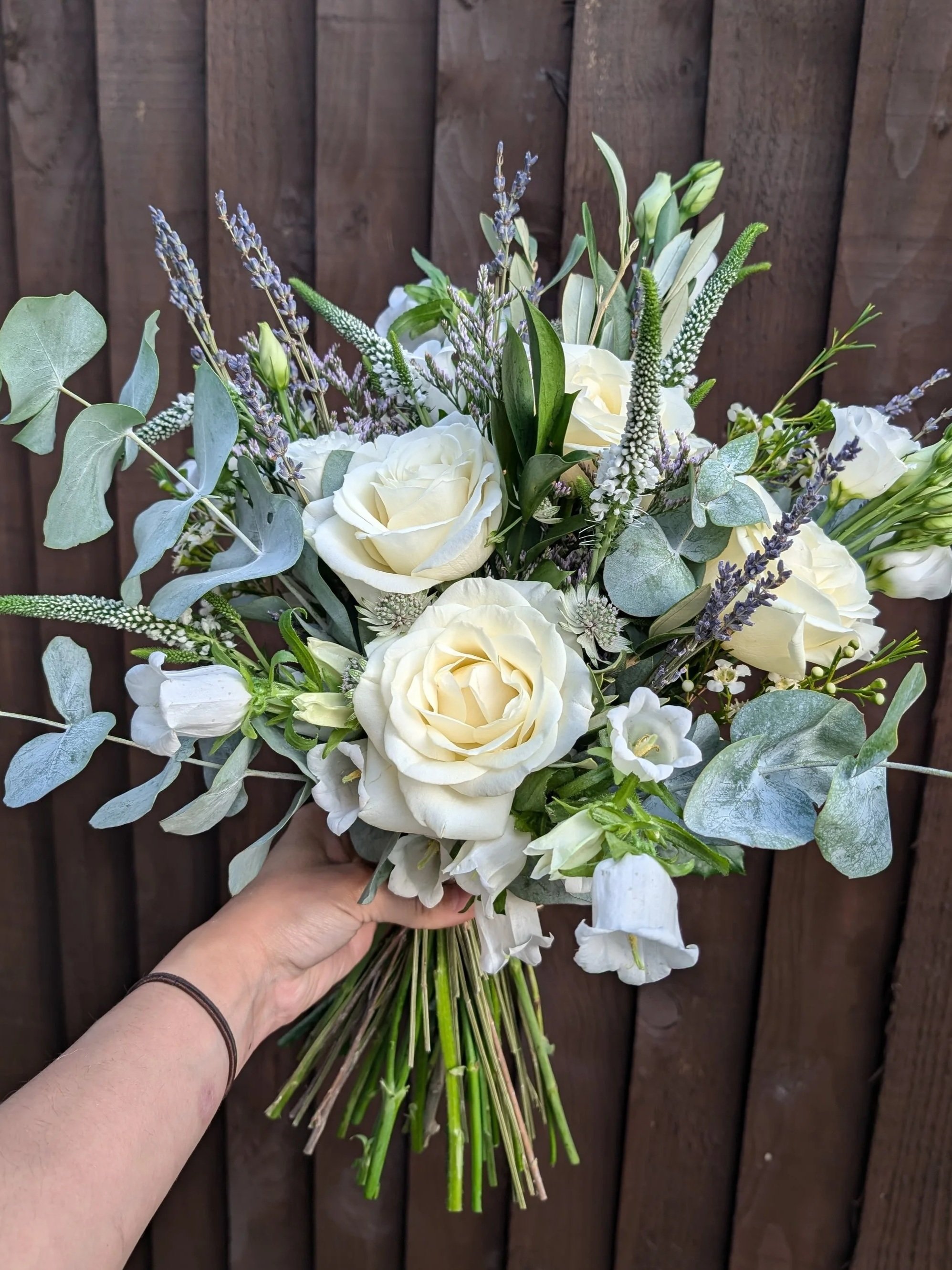 A hand holding a bouquet of white roses, lavender, and greenery in front of a wooden fence.