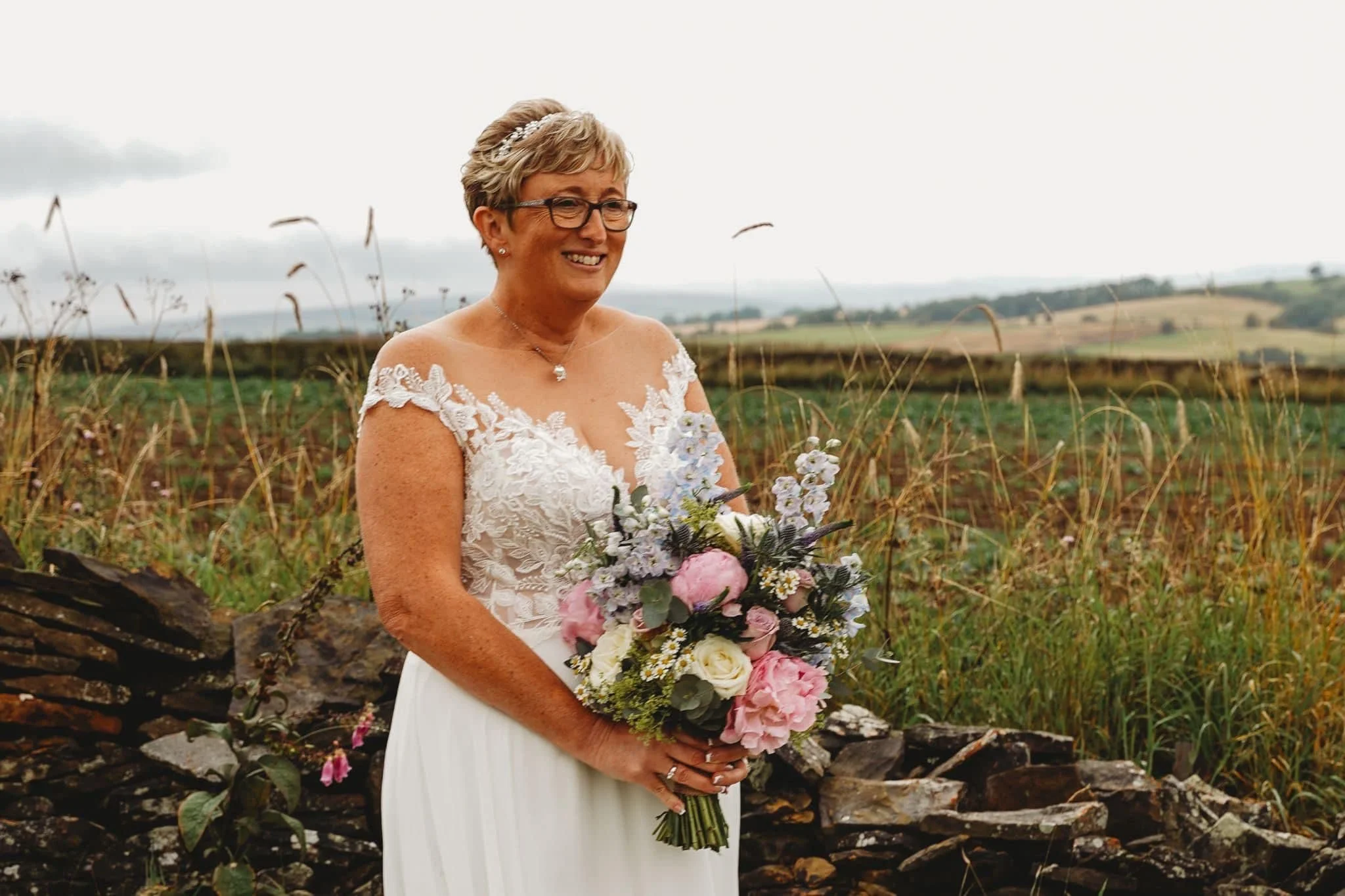 A woman in a white wedding dress holding a bouquet of pink, white, and purple flowers, standing outdoors by a stacked stone wall with green fields and a cloudy sky in the background.