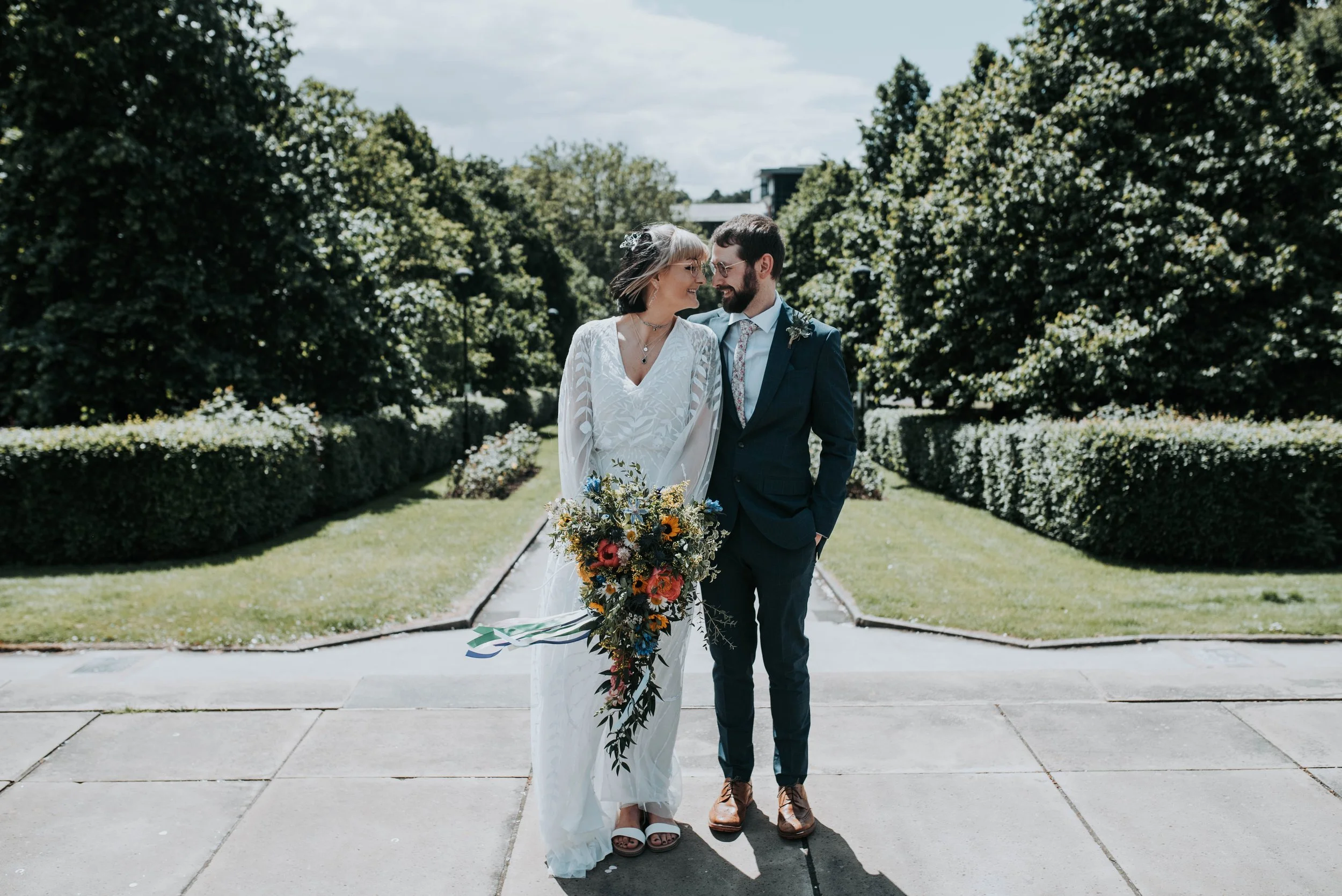A bride and groom smiling at each other outdoors on their wedding day, with lush green trees and bushes in the background.