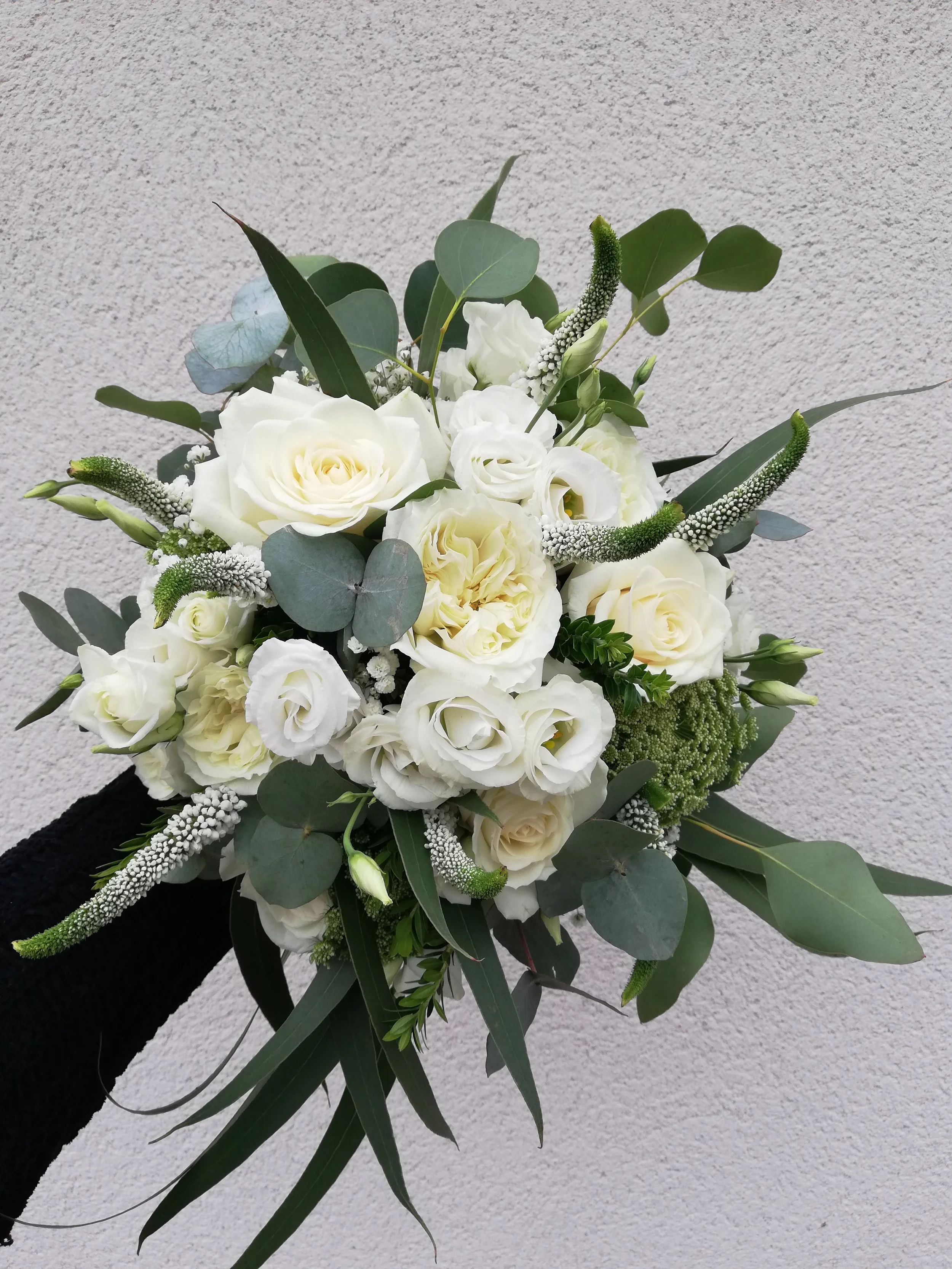 White floral bouquet with roses, lisianthus, eucalyptus, and other greenery held against a plain wall background.