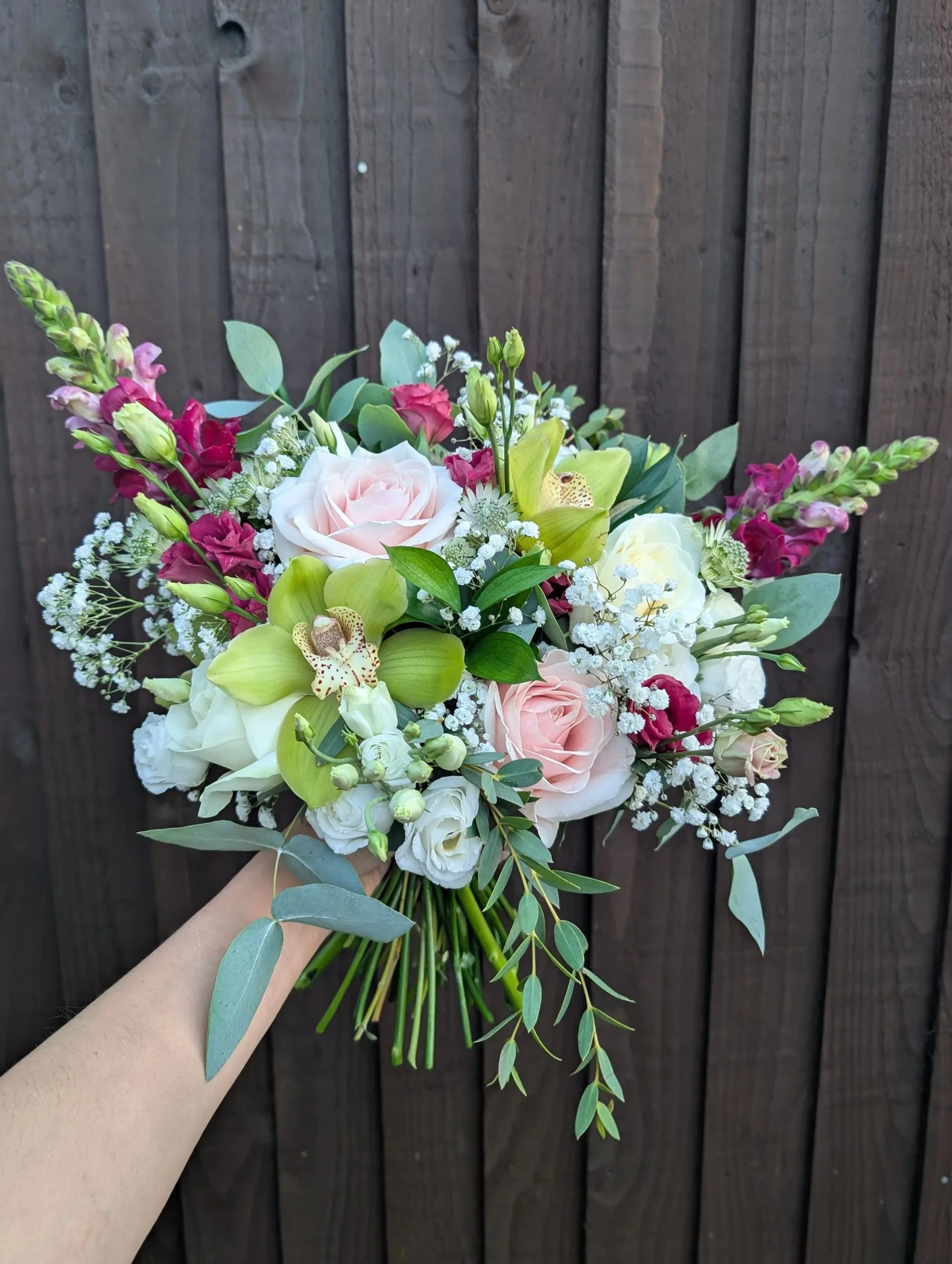 Hand holding a bouquet of pink, white, and green flowers in front of a dark wooden fence.