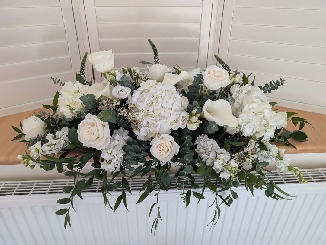 A floral arrangement with white roses, hydrangeas, calla lilies, and greenery, placed on a wooden bench against a white louvered wall.