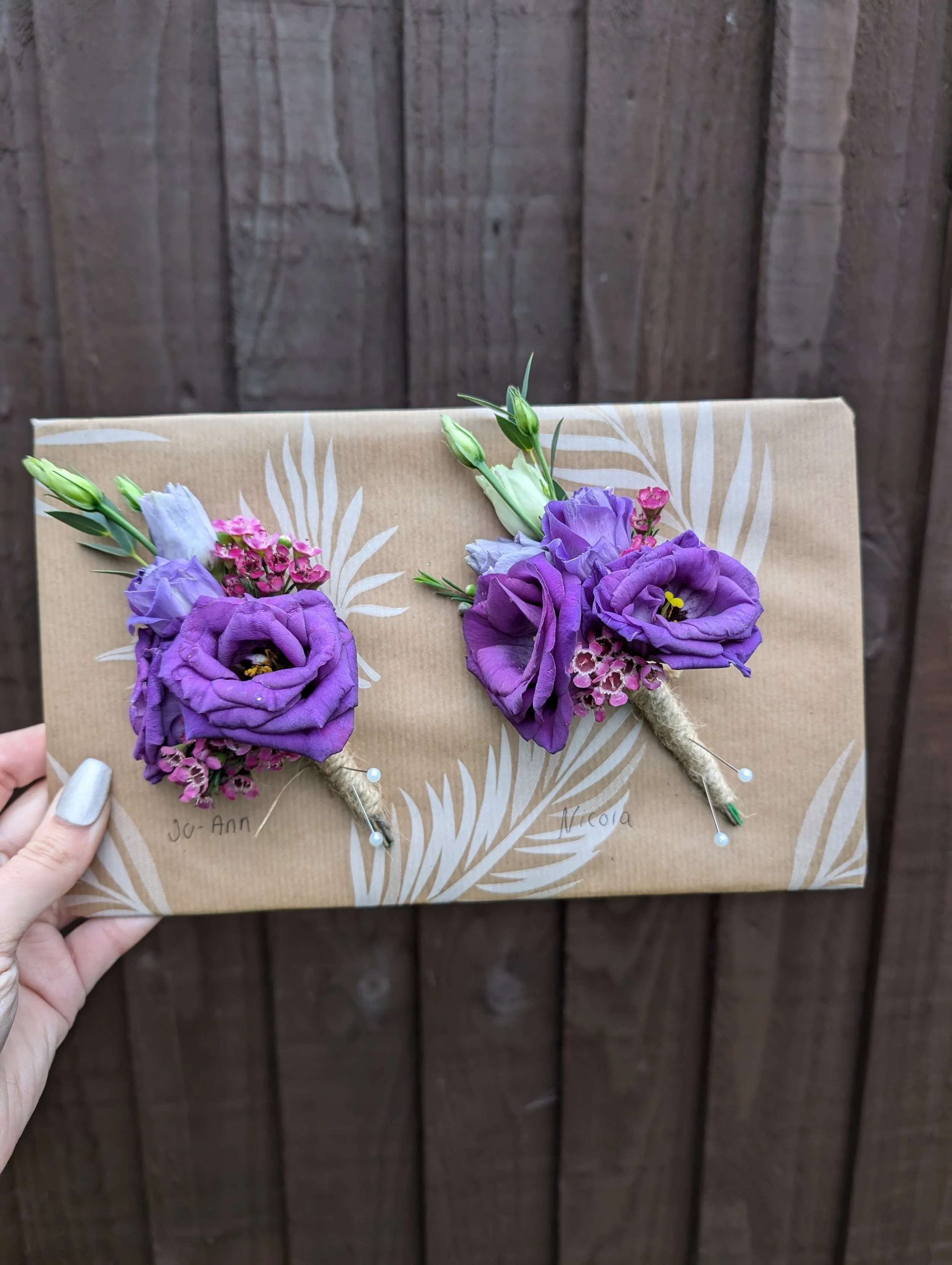 A hand holding a decorated envelope with two purple floral boutonnieres attached, labeled 'Jo-Ann' and 'Victoria', with a wooden fence background.
